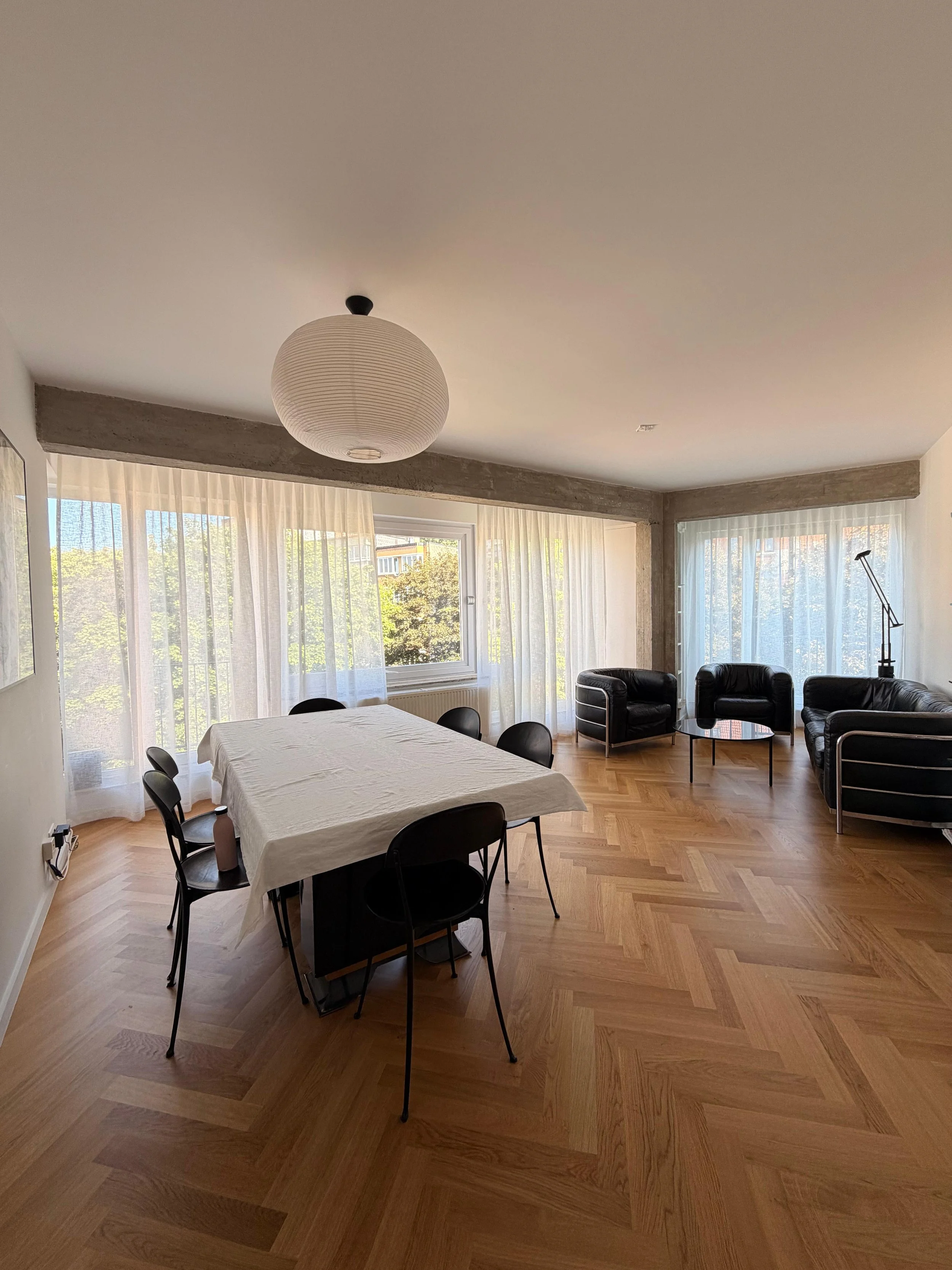 Salle à manger et salon lumineux avec fenêtres donnant sur des arbres, mobilier contemporain, tapis en bois clair, tables et chaises noires, lampadaire.