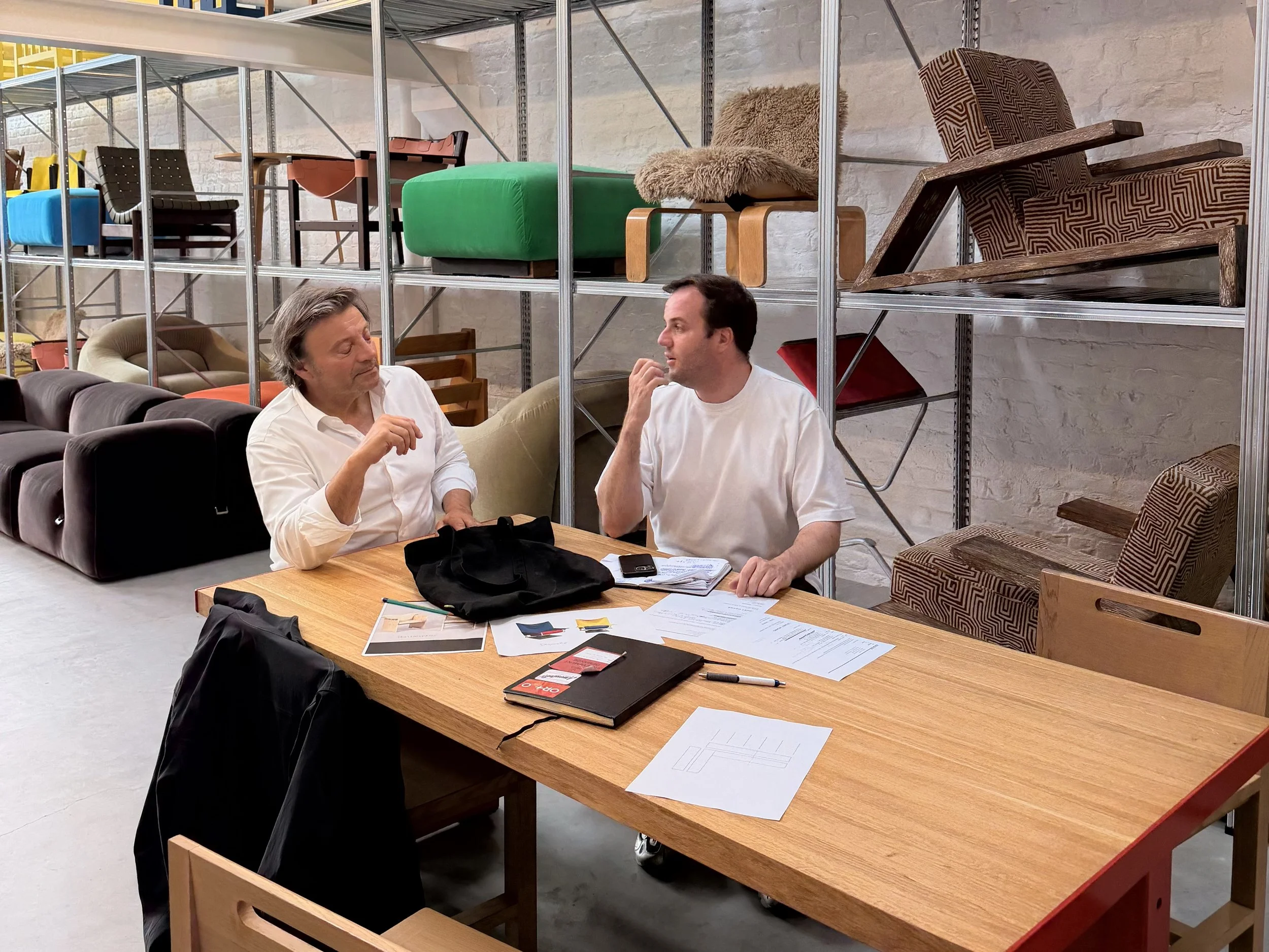 Deux hommes assis à une table en bois discutent, avec des étagères métalliques derrière eux contenant divers fauteuils et chaises.