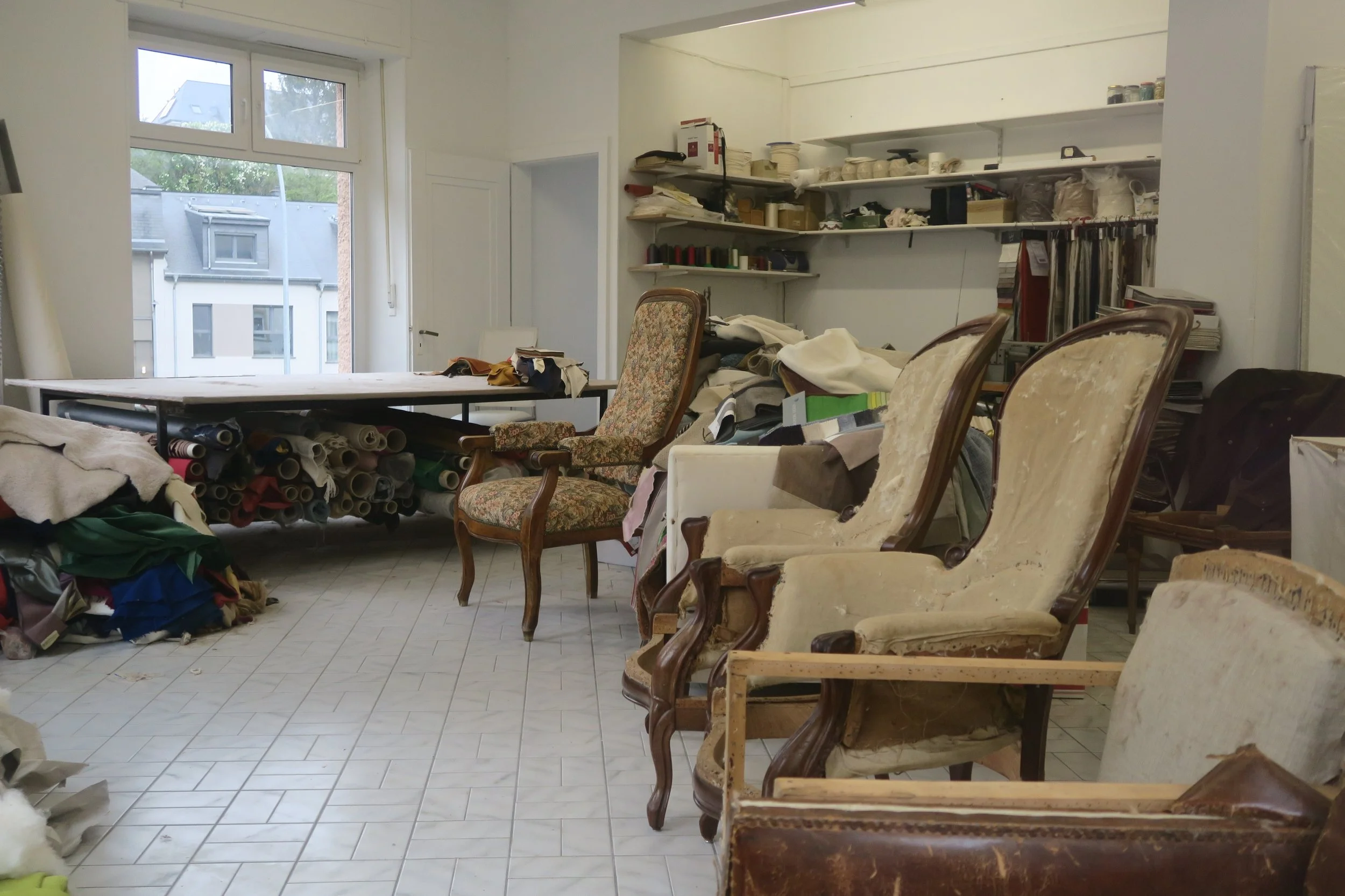 Salle encombrée avec des chaises anciennes en bois et tissu, un bureau avec des rouleaux de tissu, étagères remplies de divers matériaux, fenêtres laissant entrer la lumière naturelle.