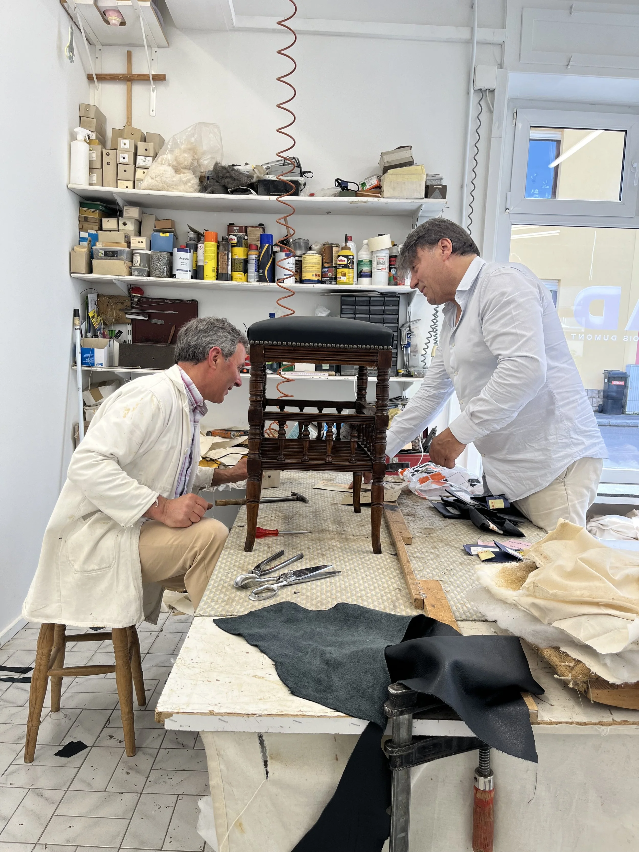 Deux hommes travaillent sur la restauration d'une chaise ancienne dans un atelier, entourés d'outils, tissus et produits chimiques. Une étagère en arrière-plan contient des boîtes, des pinceaux et des produits de réparation.