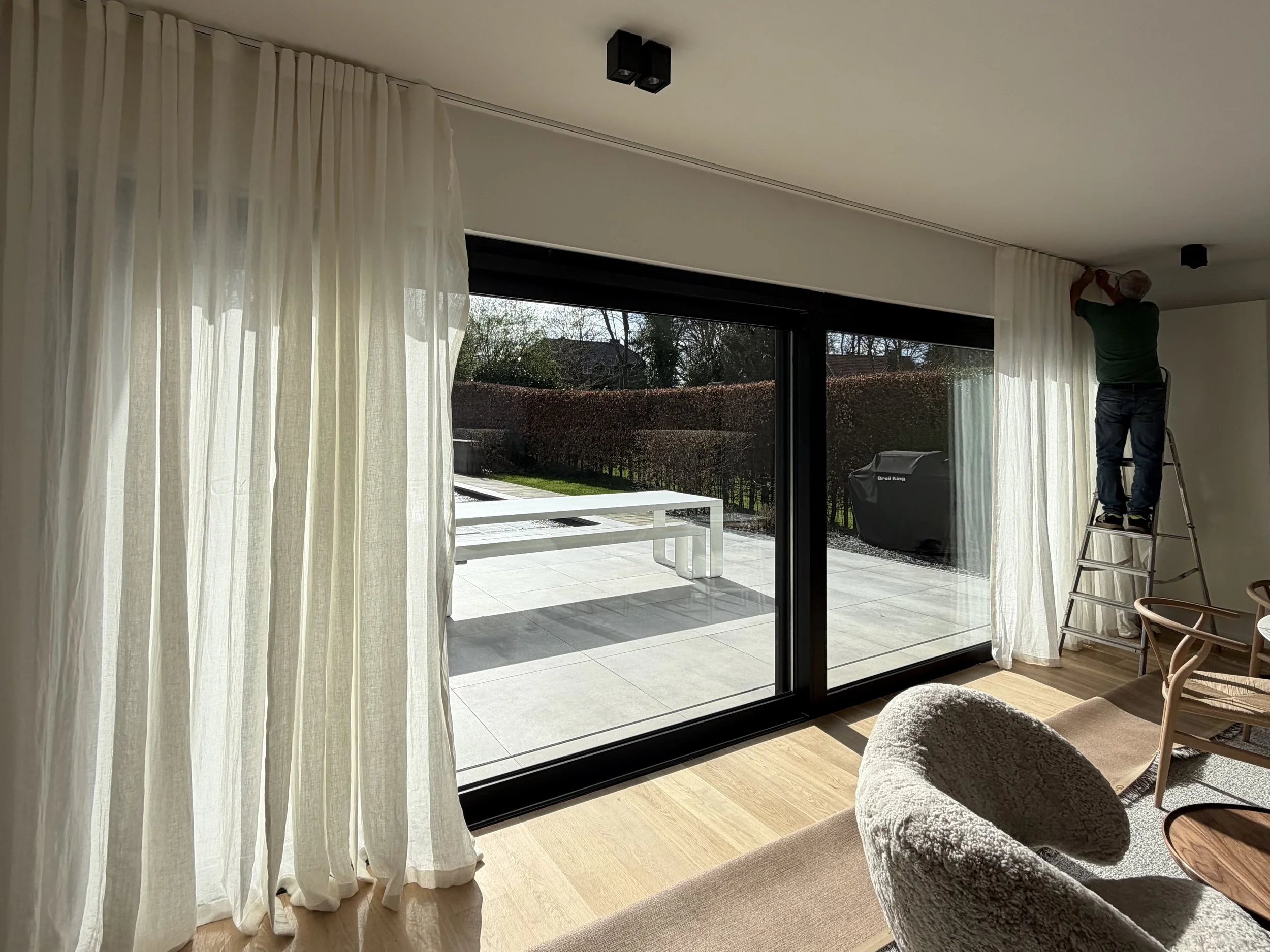 Une personne en train de poser ou d'installer une fenêtre ou une porte-fenêtre dans un salon moderne, avec des rideaux blancs et un espace extérieur visible.