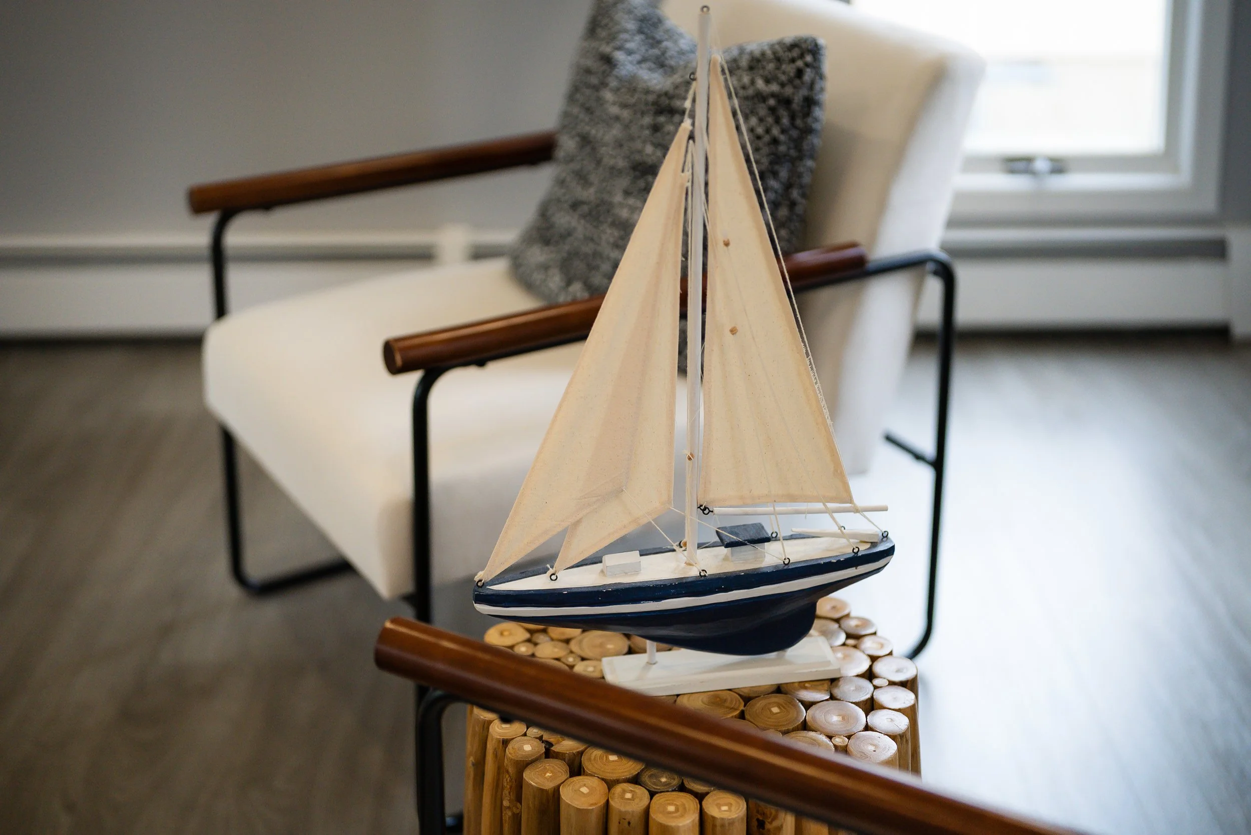 A decorative model of a sailboat on a wooden stand, placed on top of a round wooden table with circular wooden slices underneath, with a white couch and gray pillow in the background near a window.