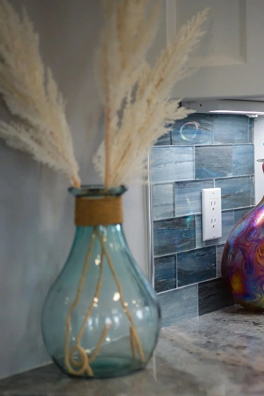 Close-up of a glass vase with dried pampas grass on a kitchen countertop, with a mosaic tile backsplash and a decorative colorful vase in the background.