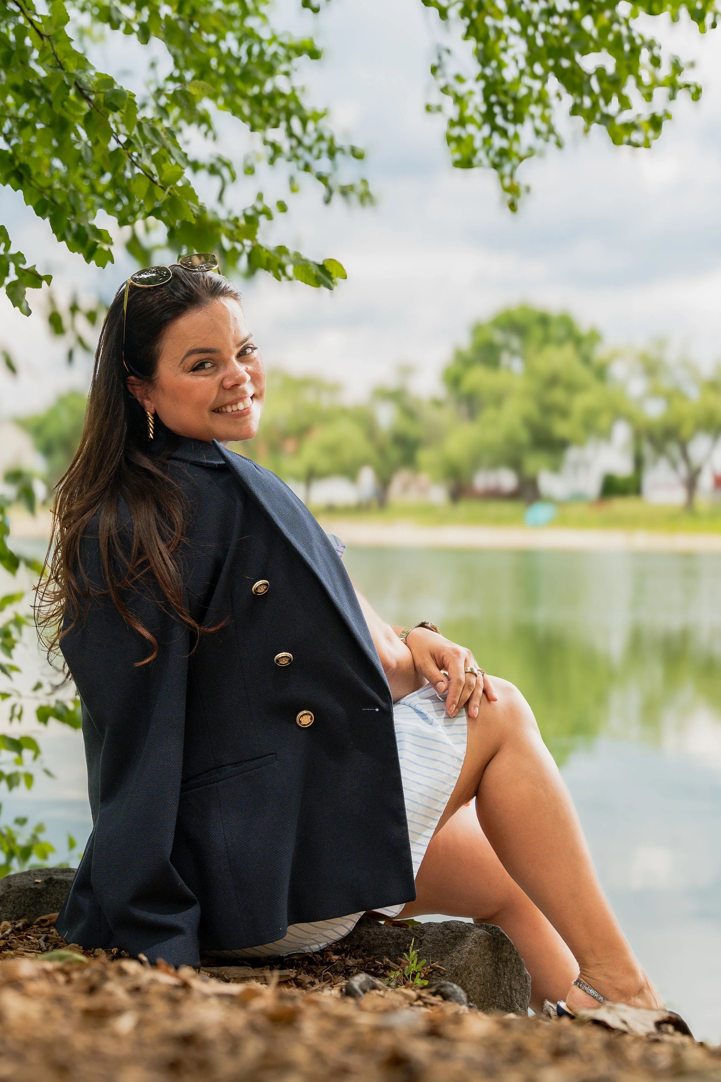 A woman sits outdoors beside a lake, smiling at the camera. She has long dark hair, is dressed in a black blazer and sunglasses, with a backdrop of trees and a cloudy sky.