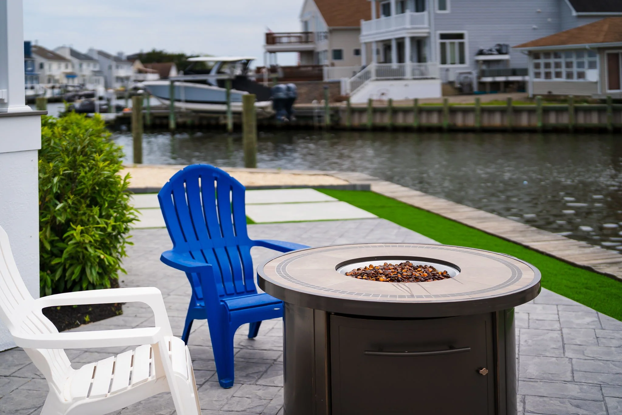 A patio with a round table filled with cereal, surrounded by white and blue chairs, overlooking a waterway with boats and houses in the background.