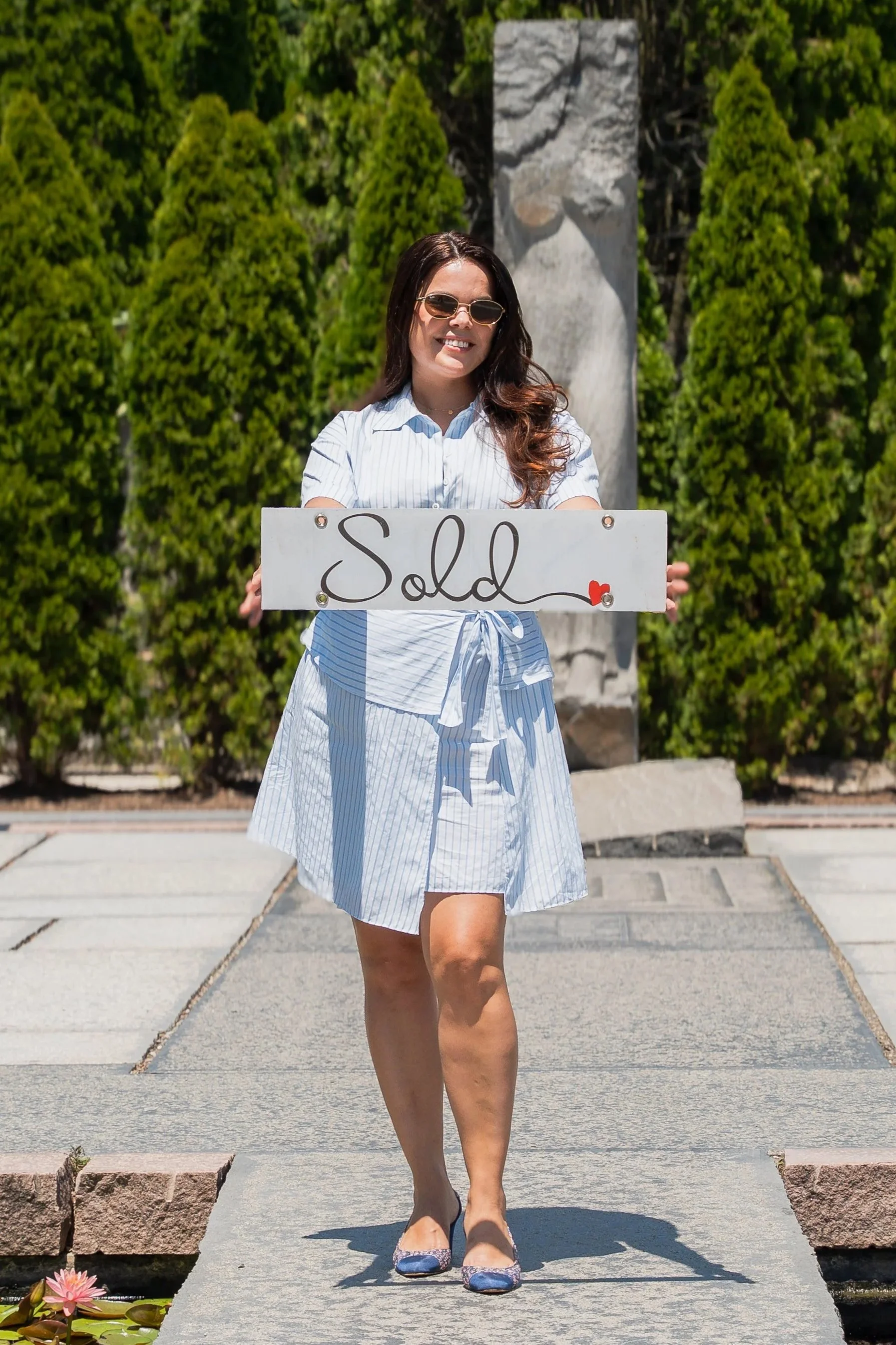 Woman with long brown hair wearing sunglasses, a light blue and white striped dress, and floral shoes, holding a sign that says 'Sold' with a small red heart, standing on a stone walkway in front of greenery and a stone monument.