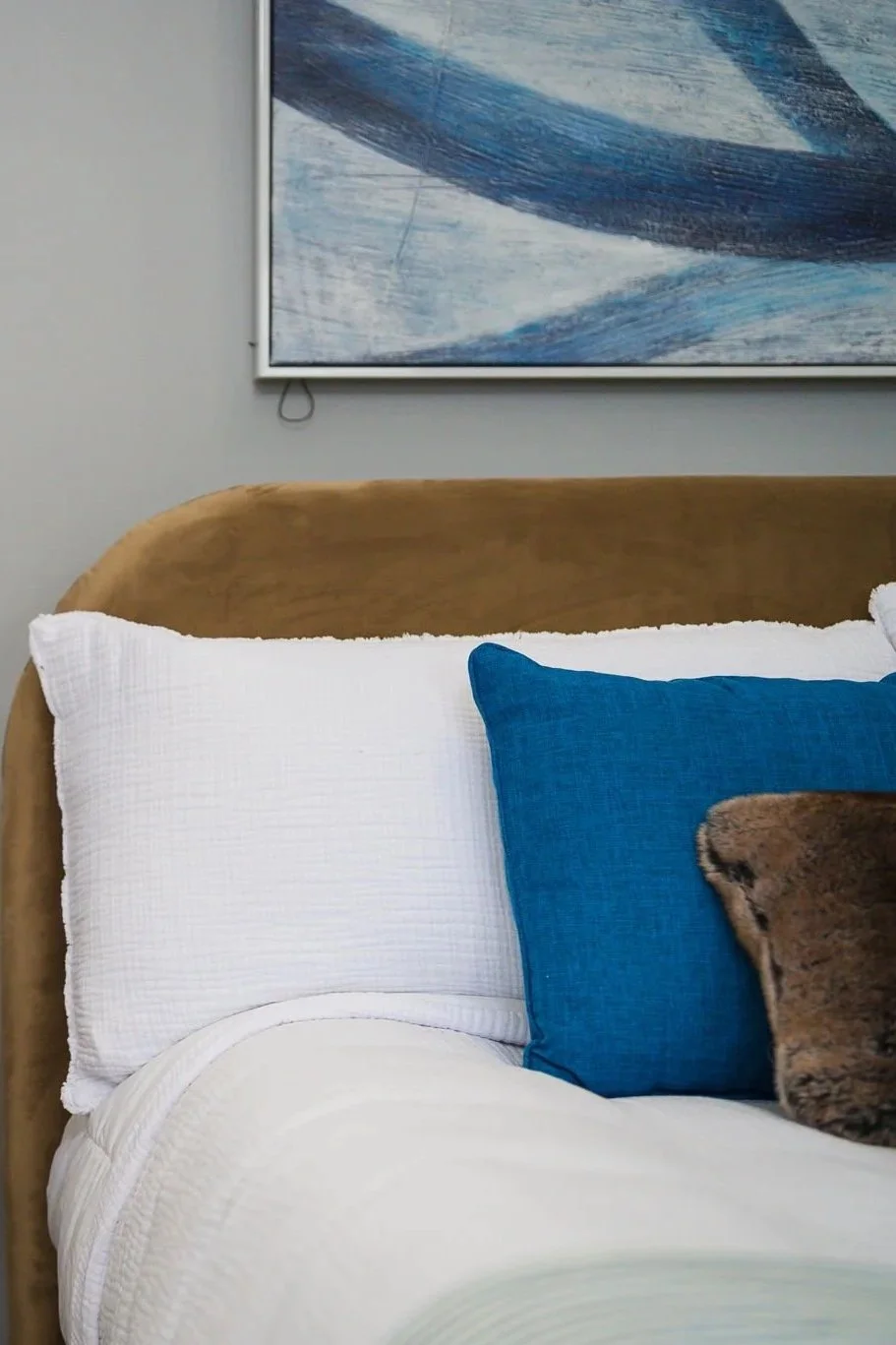 Close-up of a bed with white and blue pillows, a brown headboard, and an abstract blue and white painting on the wall.