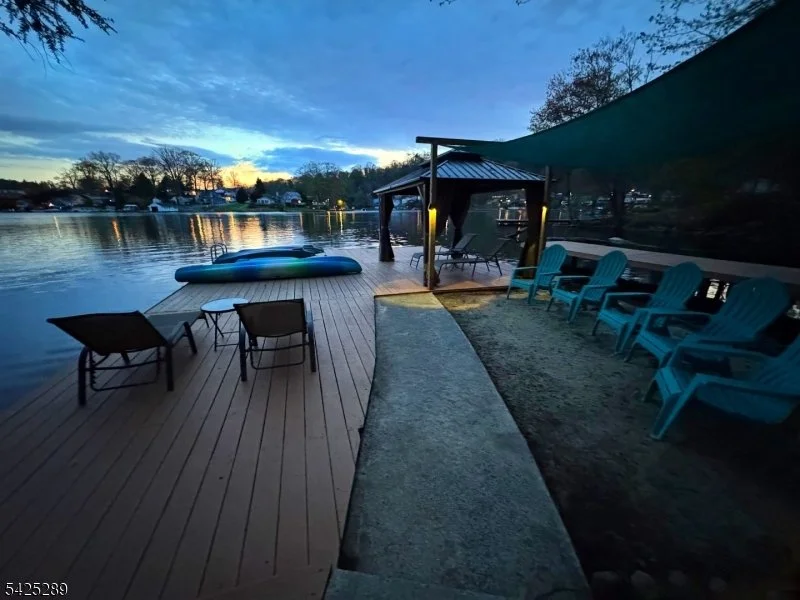 A lakeside scene at sunset with a wooden deck, lounge chairs, and a covered seating area with additional chairs under a shade sail. The calm lake reflects the sky, surrounded by trees and houses.