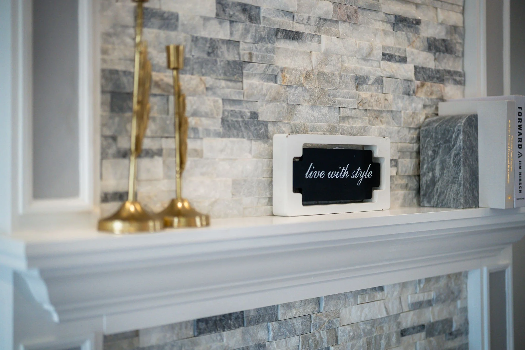 Decorative interior shelf with gold candlesticks, a framed quote reading "live with style," a gray marble book, and a stone accent wall.