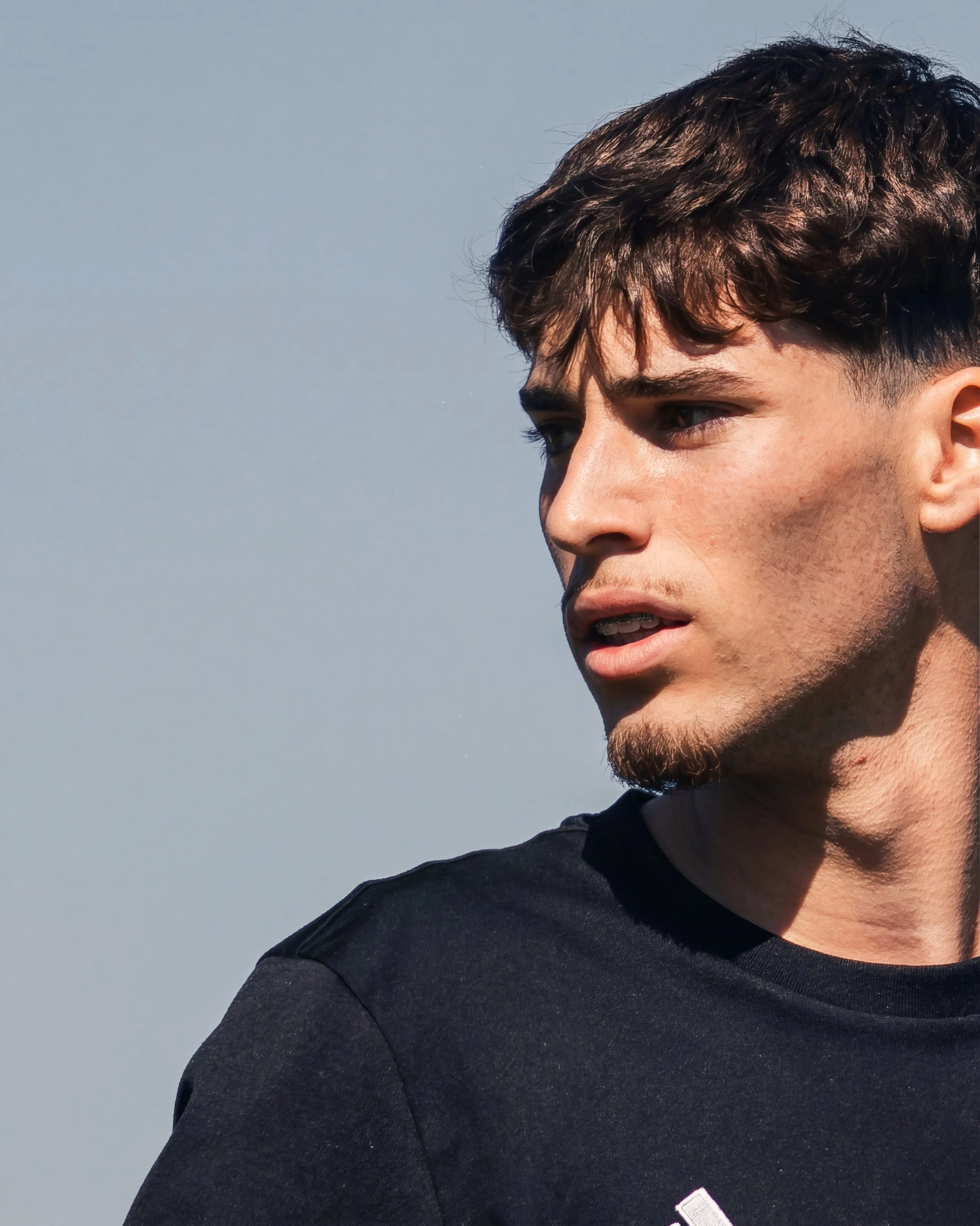 Close-up of a young man's face and upper body, with short dark hair, wearing a black shirt, outdoors against a light blue sky.