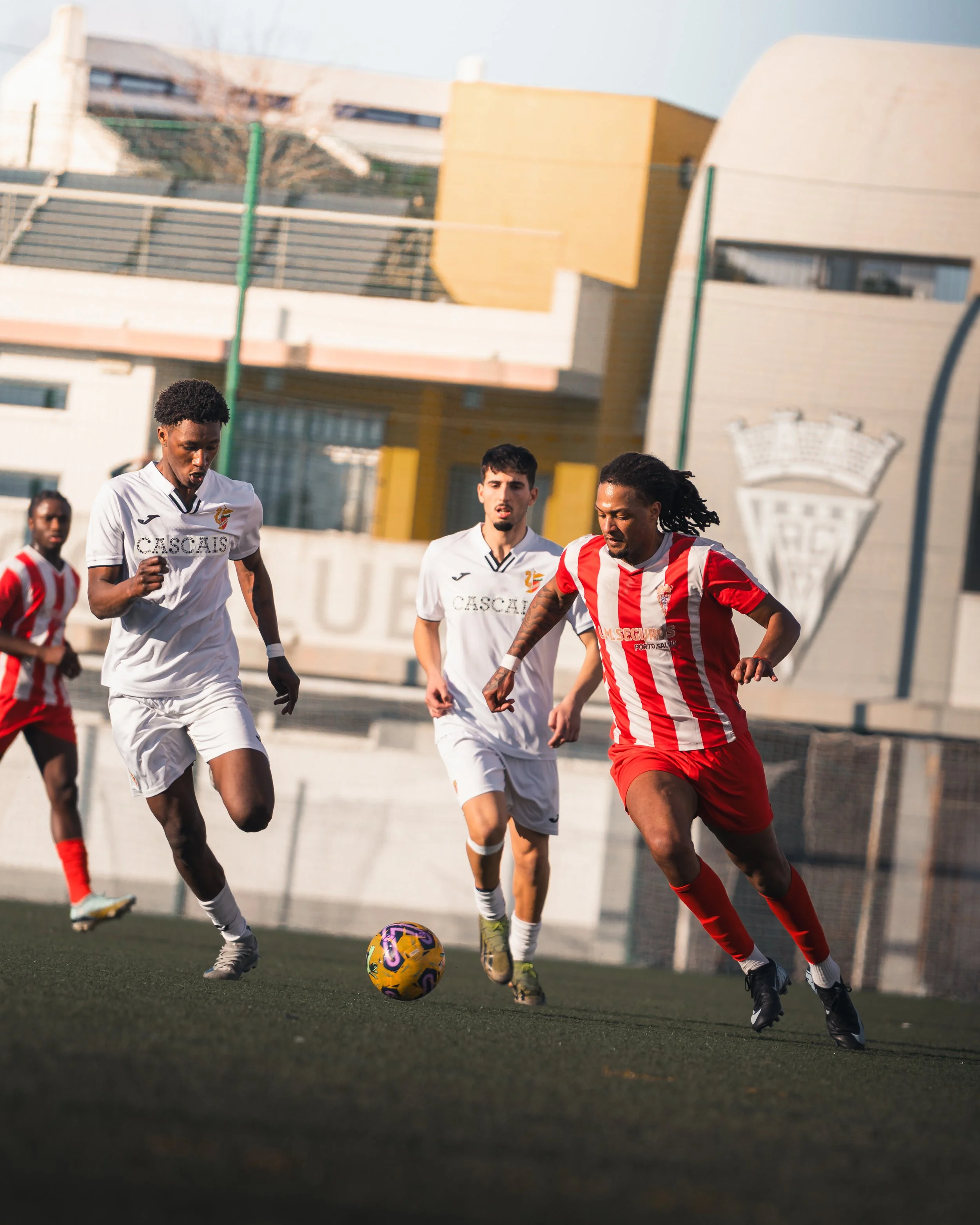Four football players on the field, two wearing white jerseys and two wearing red and white striped jerseys, racing after a yellow and purple soccer ball during a game.