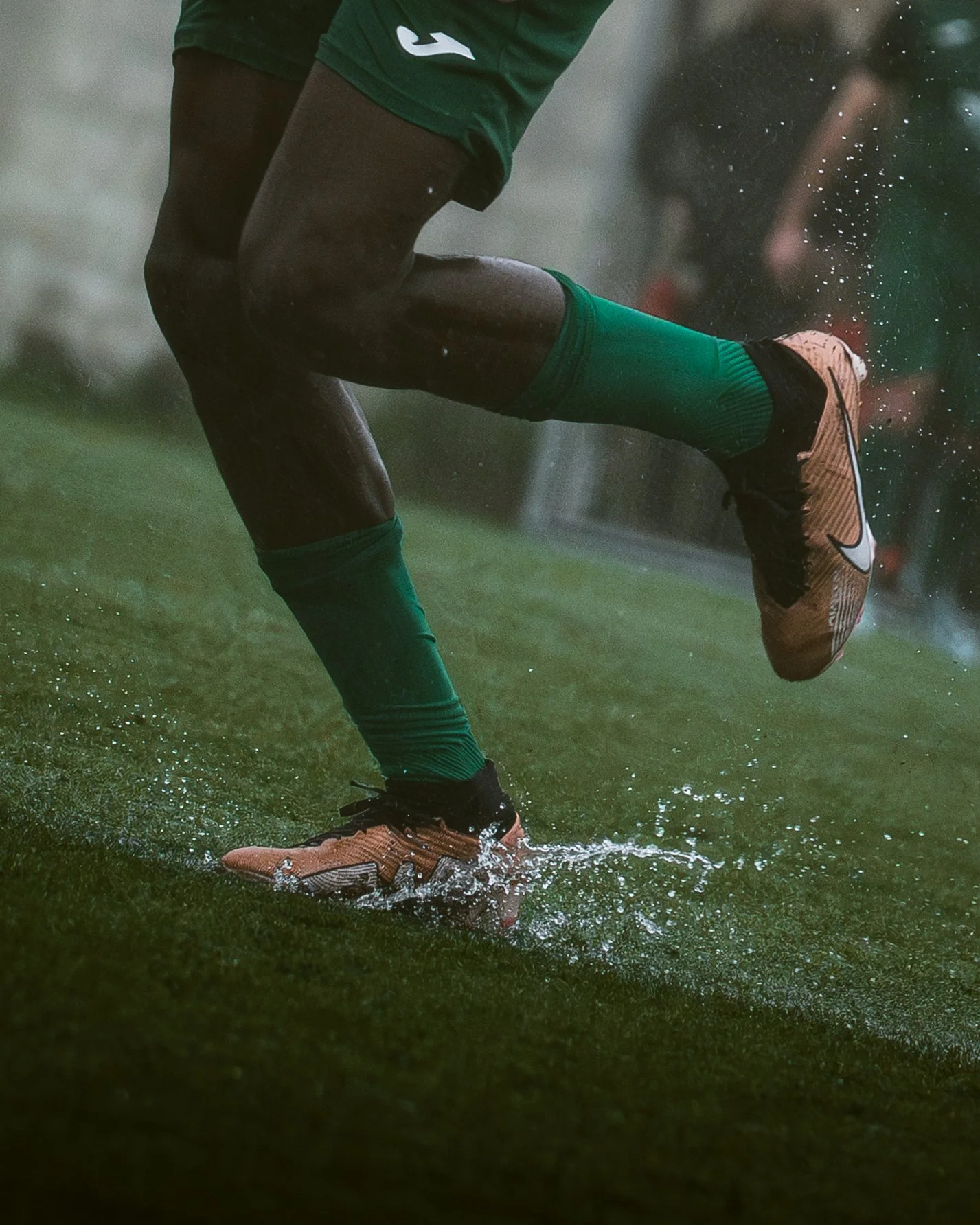 Close-up of a football player's legs and feet as they kick a ball on a wet field, wearing green socks and bronze cleats.