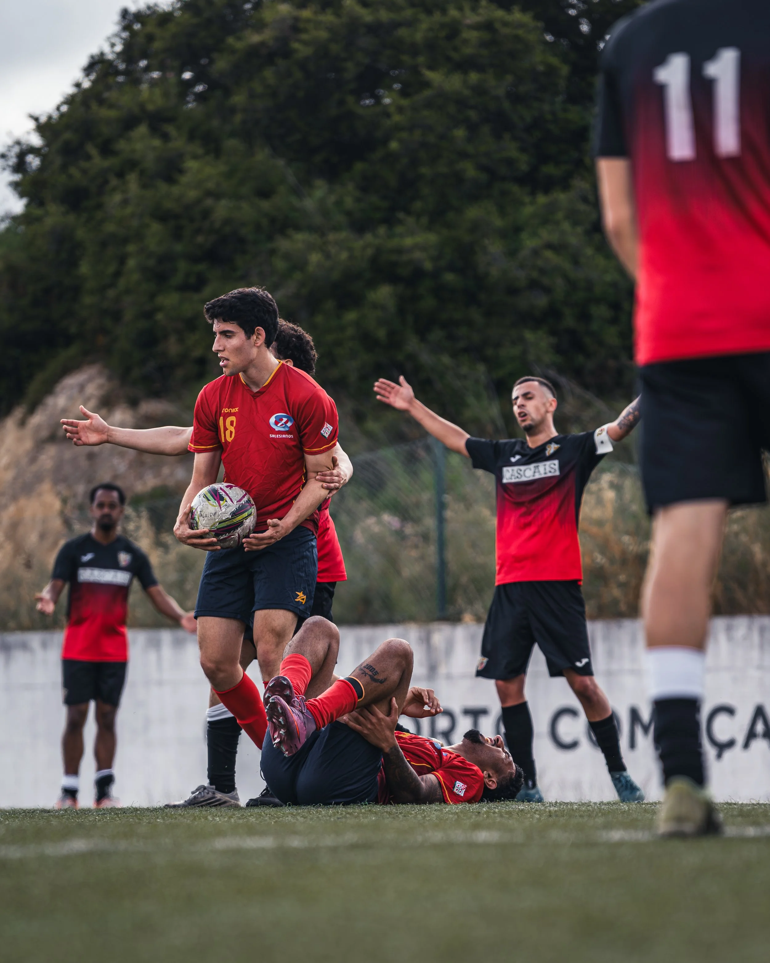 Football players on the field during a match, with one player from the team in red holding a soccer ball and standing over a fallen opponent, while other players and officials are visible in the background.