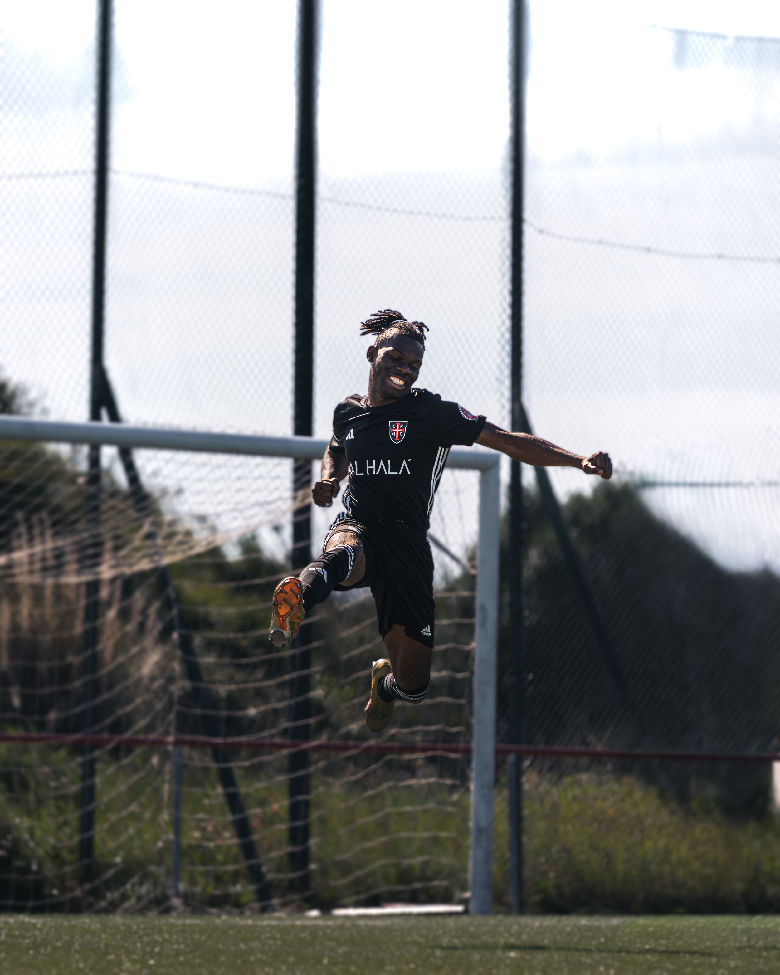 A football player in black uniform is celebrating a goal on a soccer field, with a chain-link fence and a cloudy sky in the background.