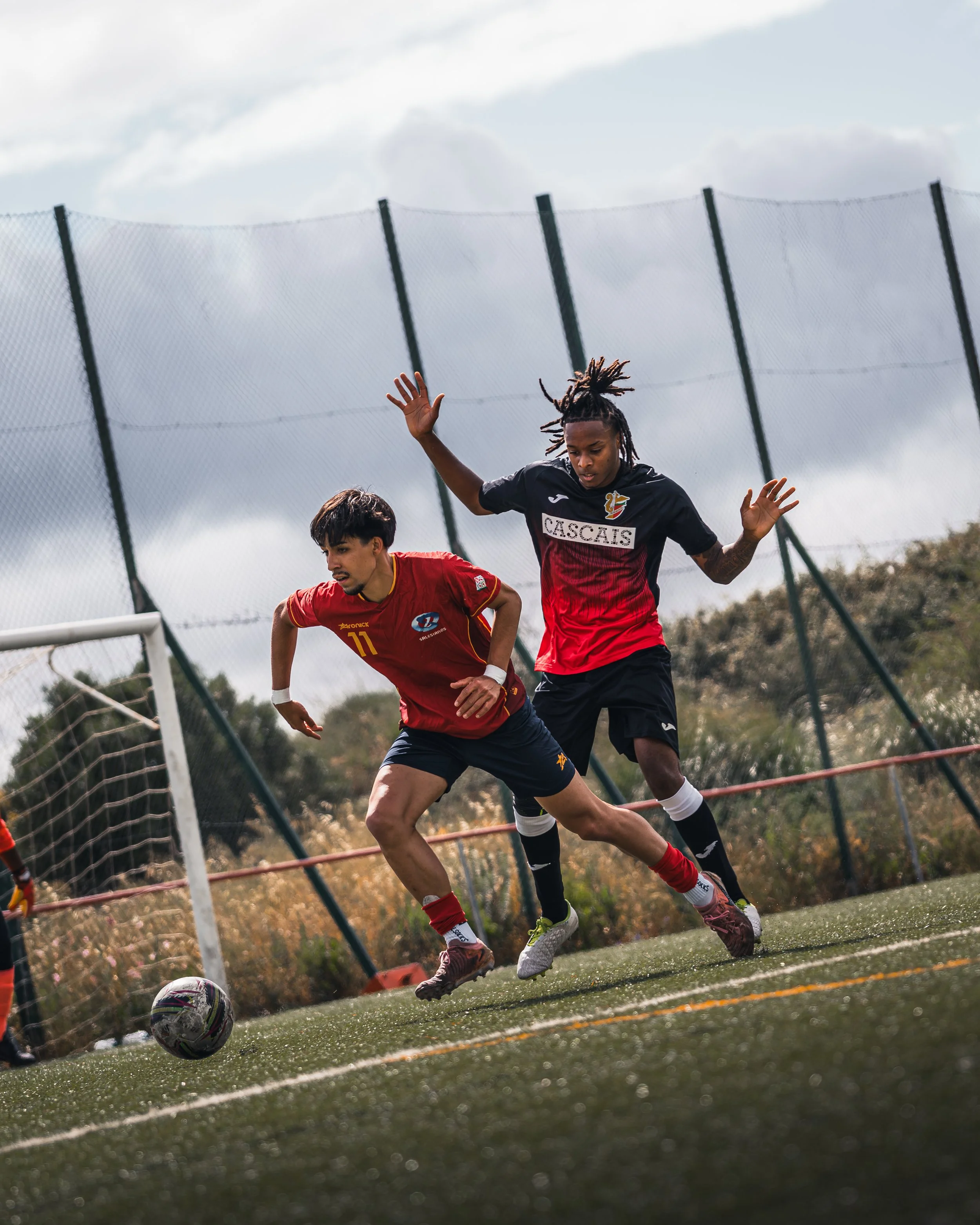 Two male soccer players compete for the ball on a field near a goal, with a fence and cloudy sky in the background.