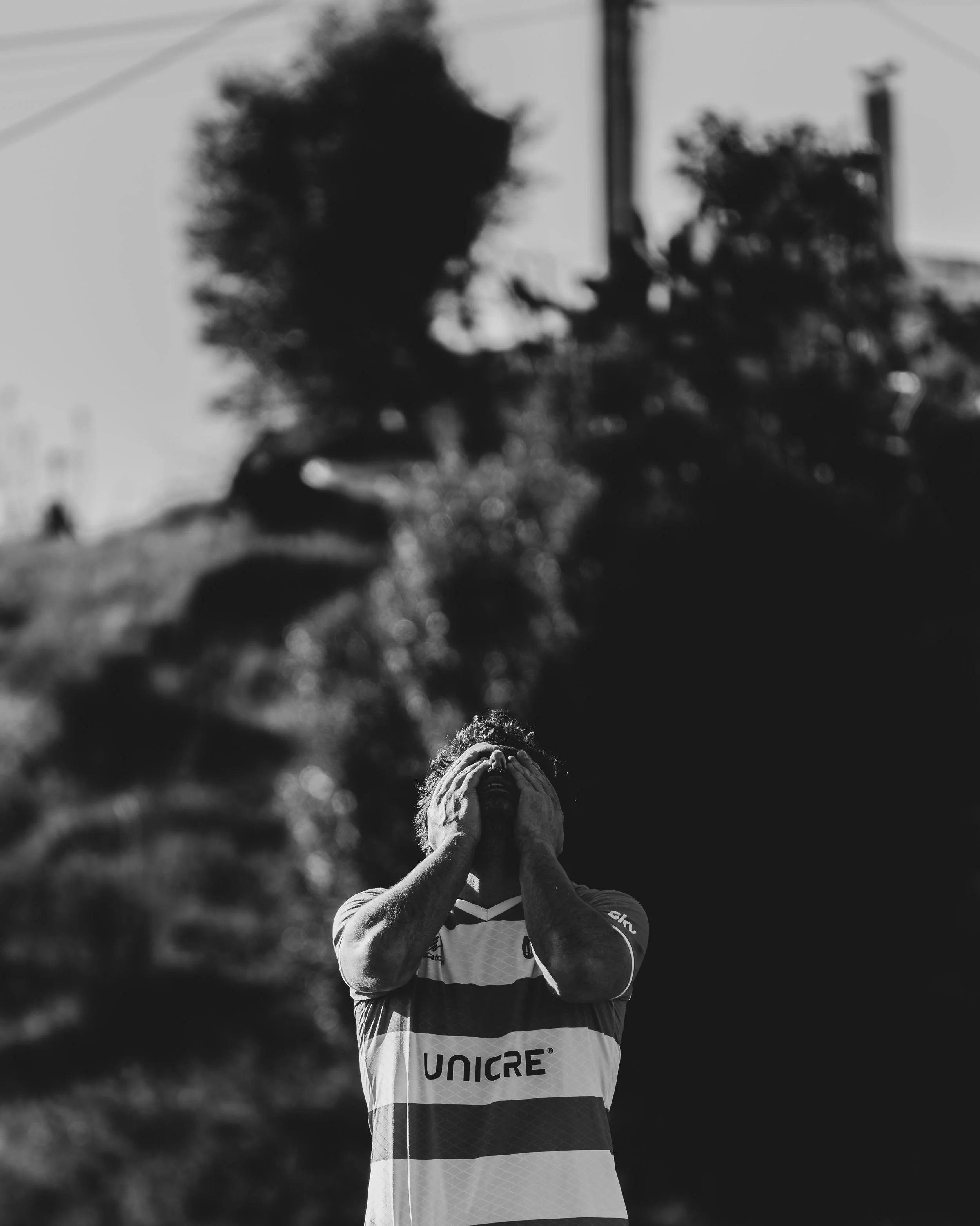 A person wearing a sports jersey with the word 'UNICRE' on it, covering their face with their hands, standing outdoors in front of blurred trees and power lines.