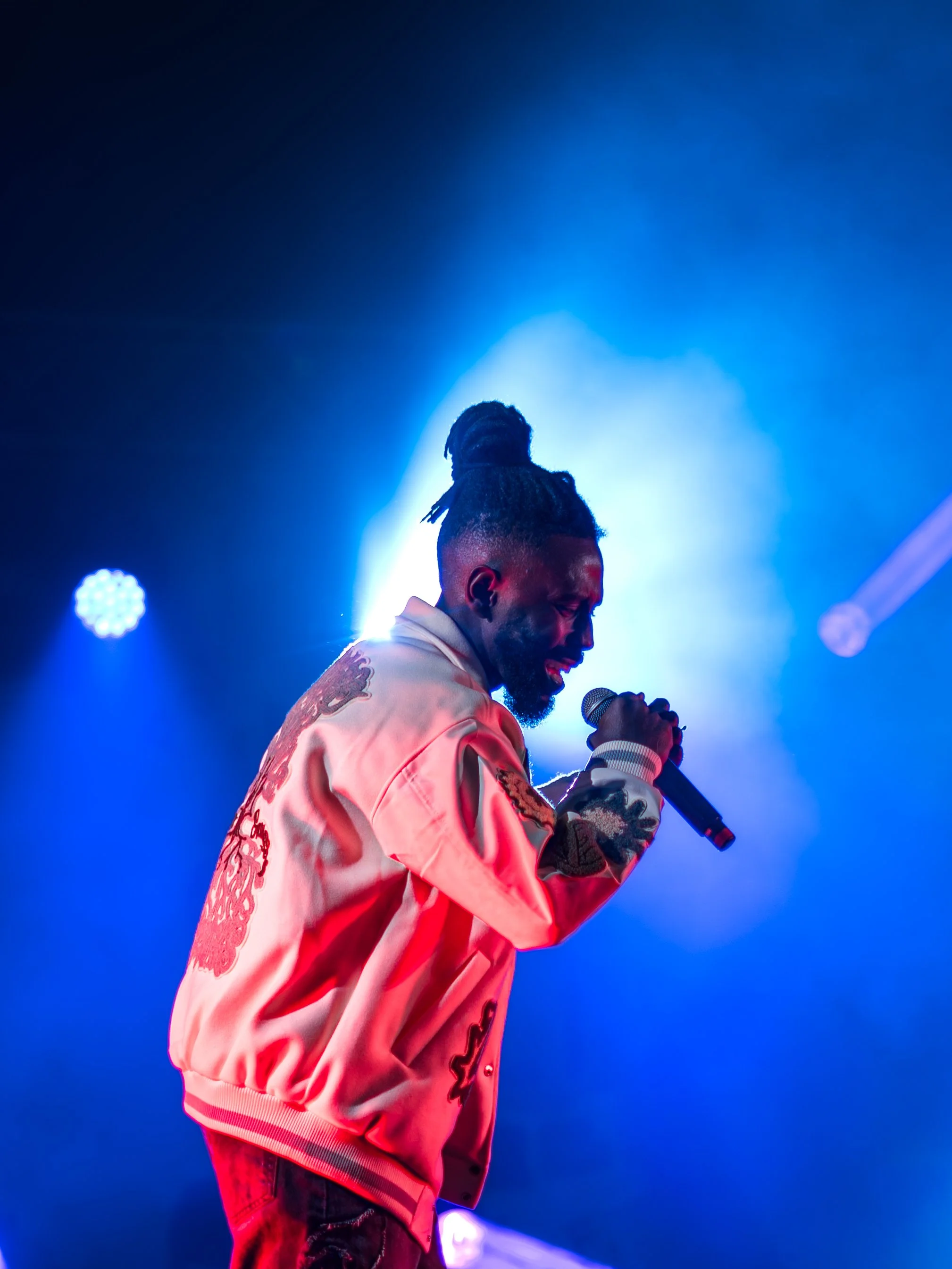 A male performer singing into a microphone on stage, with bright blue stage lighting and wearing a pink satin jacket with embroidery and dark jeans.