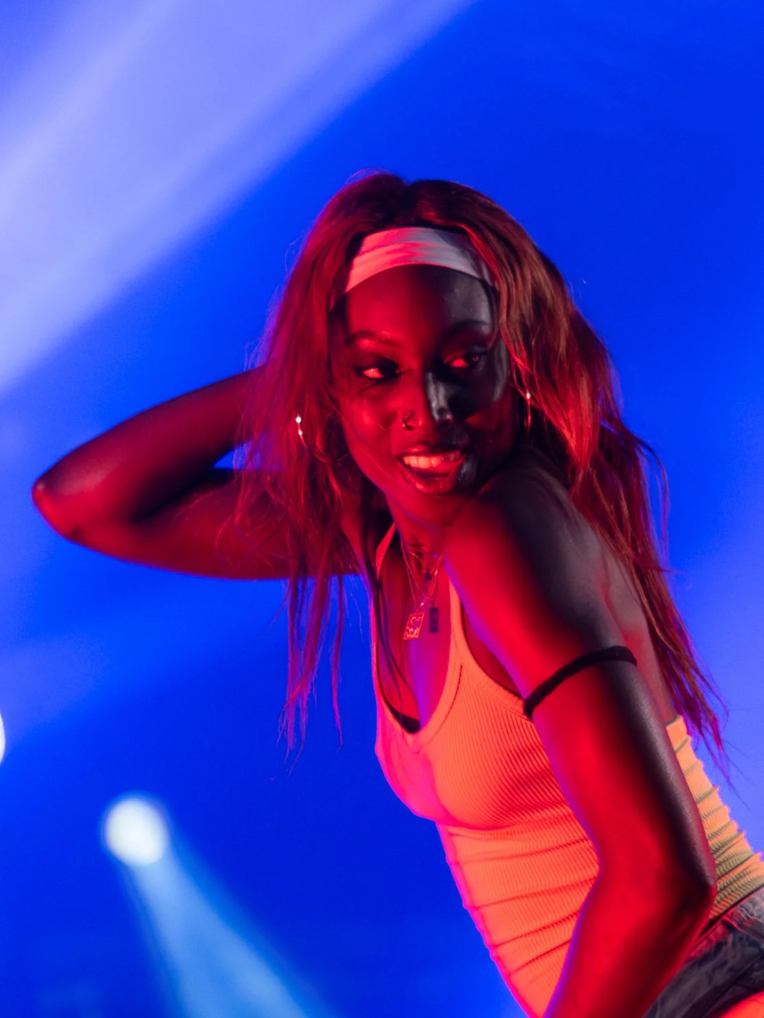 A young woman with red hair and a white headband posing against a blue background with pink and purple lighting.