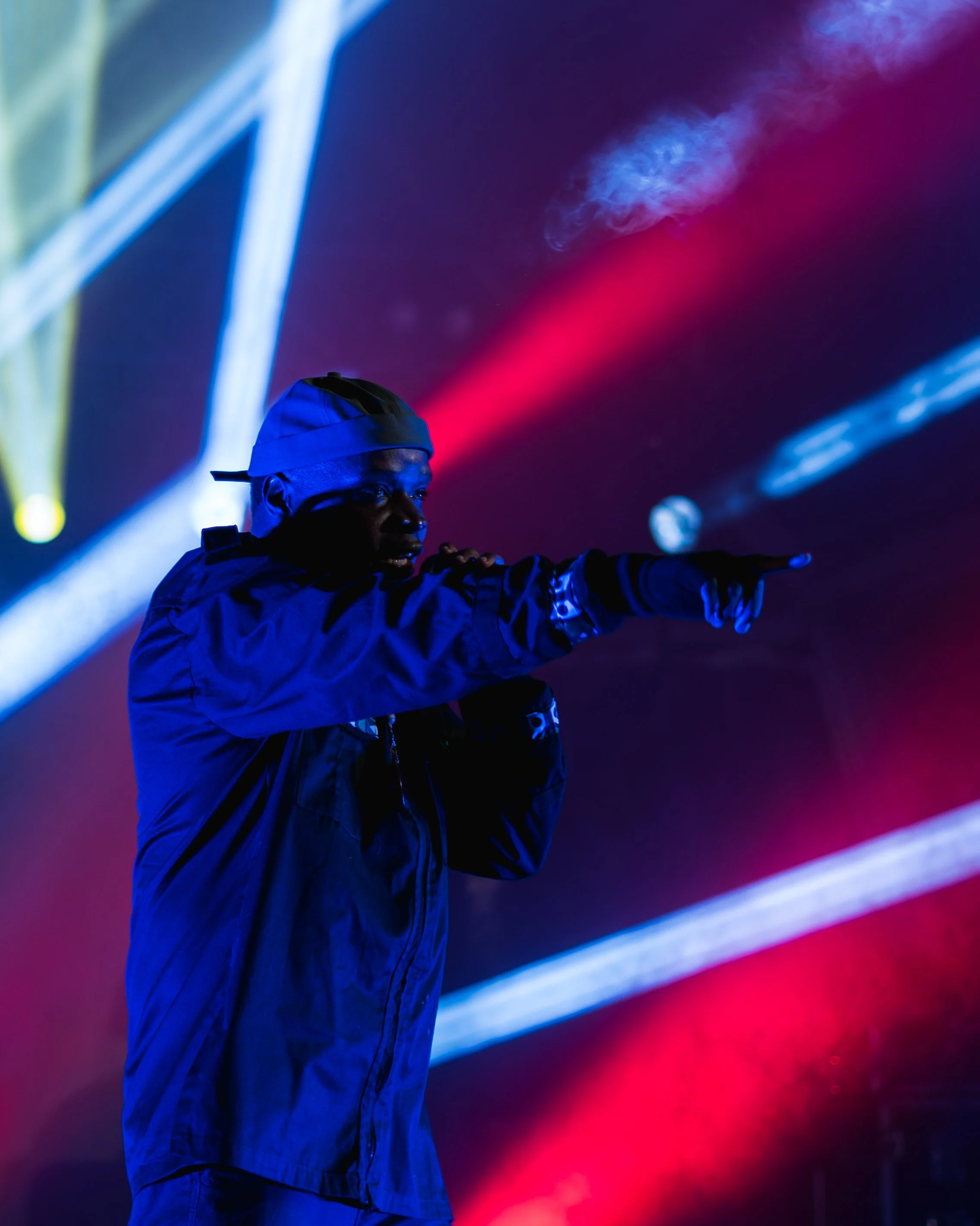 A person in dark clothing and a white cap is standing against a vibrant background of red, blue, and purple light streaks, pointing towards the right.