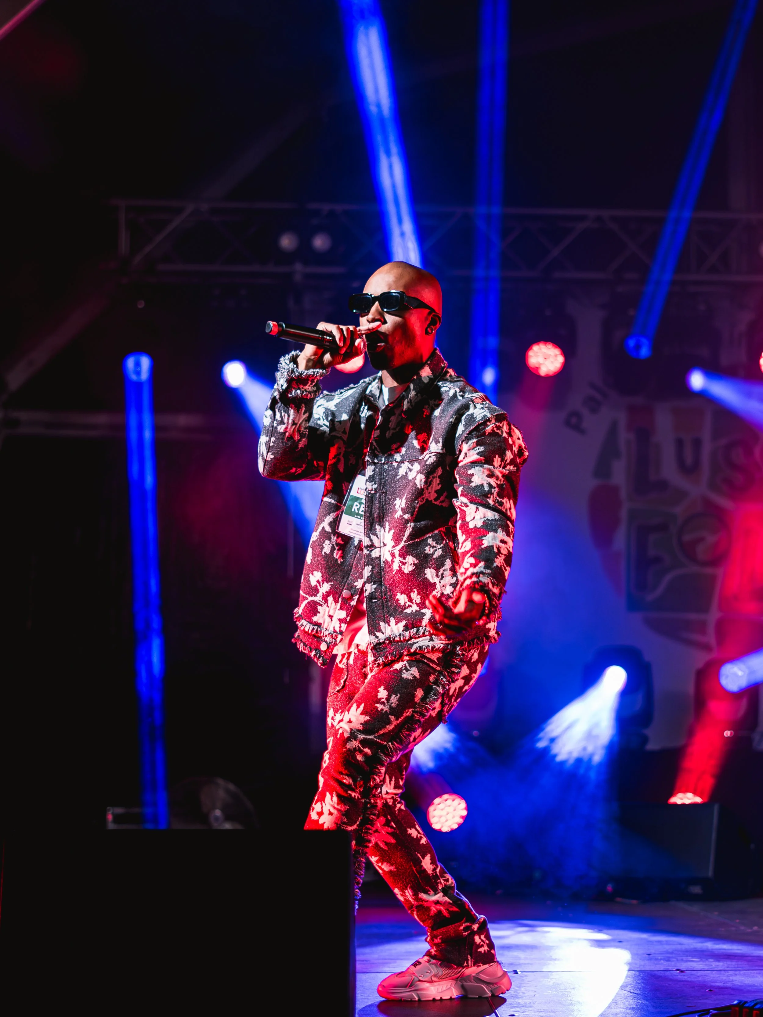 A rapper wearing sunglasses, a camo print jacket, and matching pants, performing on stage with colorful lighting effects.