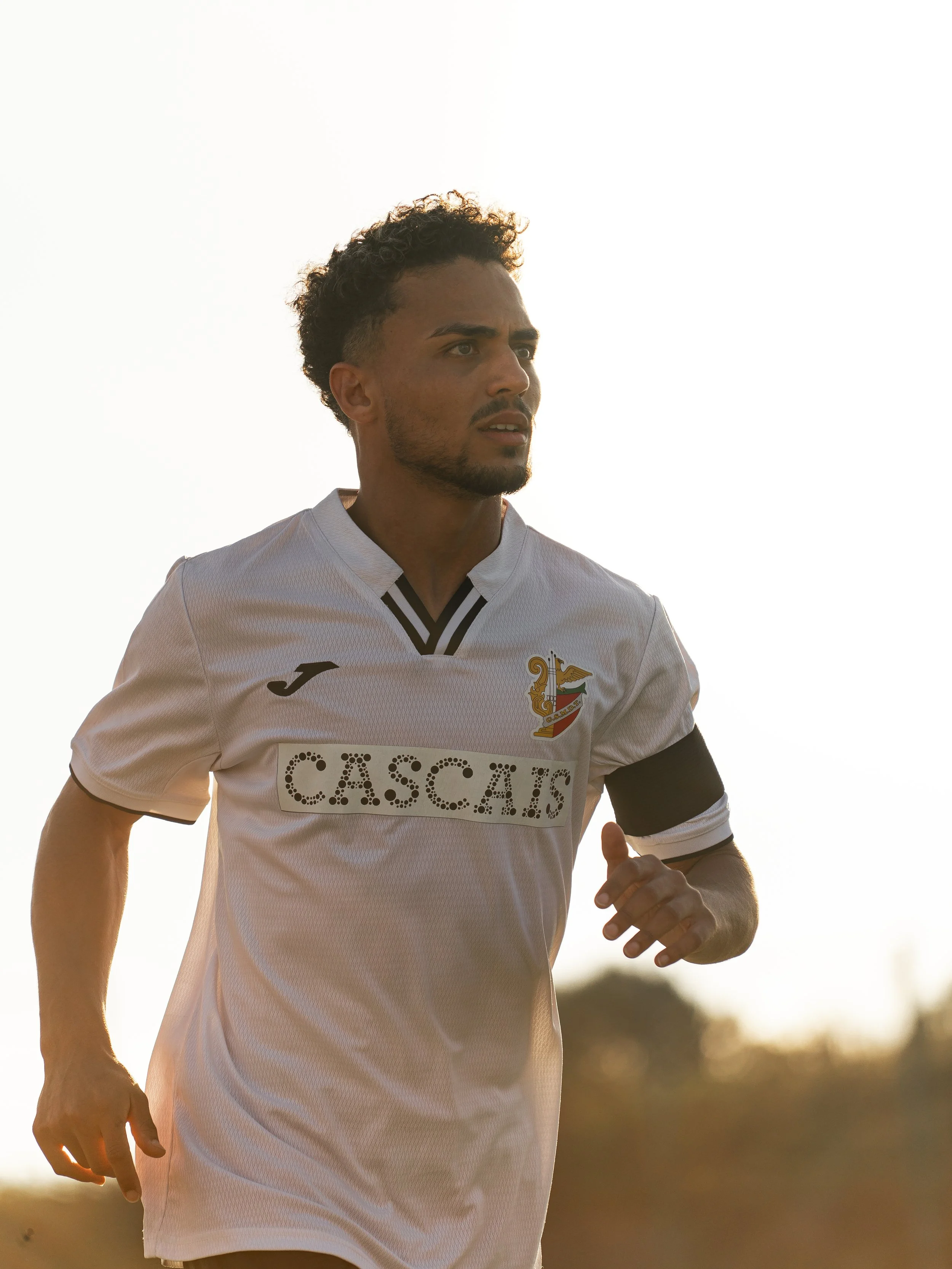 A male football player is running outdoors during sunset. He is wearing a white jersey. His expression is focused and determined, with curly hair and a beard.
