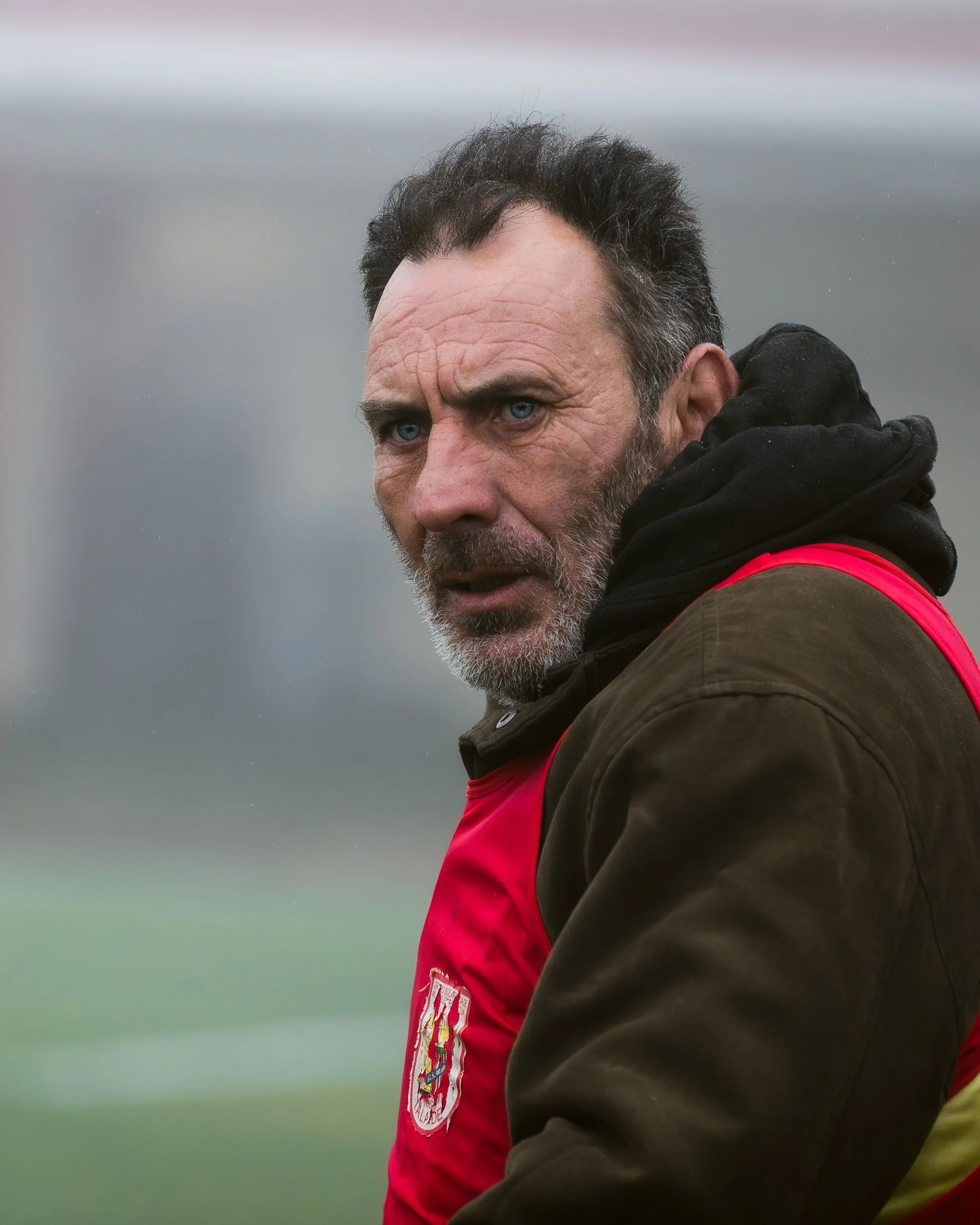 Close-up of a man with dark hair, blue eyes, and gray beard, wearing a black hoodie and a red vest, outdoors on a foggy day.