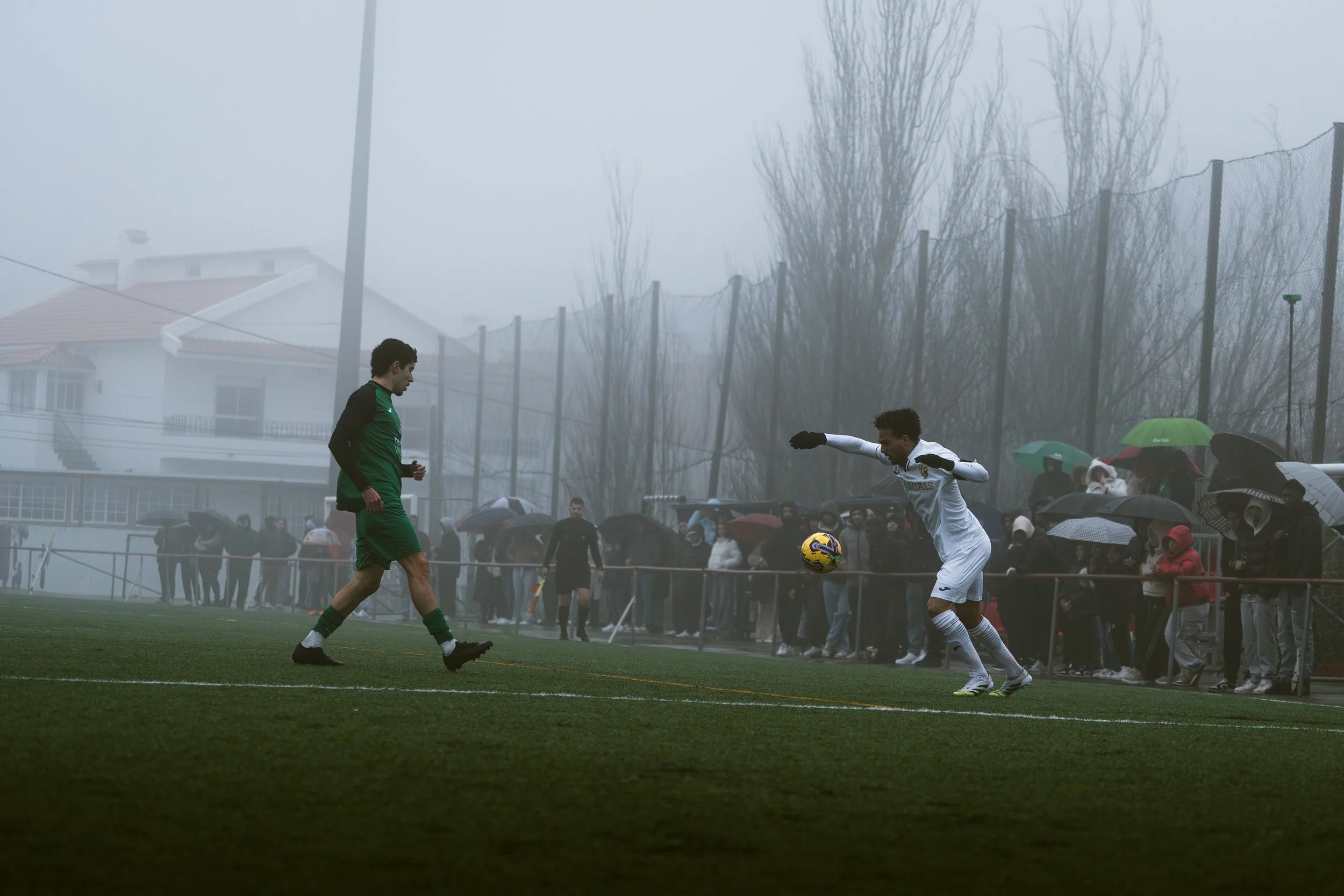 Two football players are competing for the ball on a rainy day, with a crowd of spectators holding umbrellas in the background.