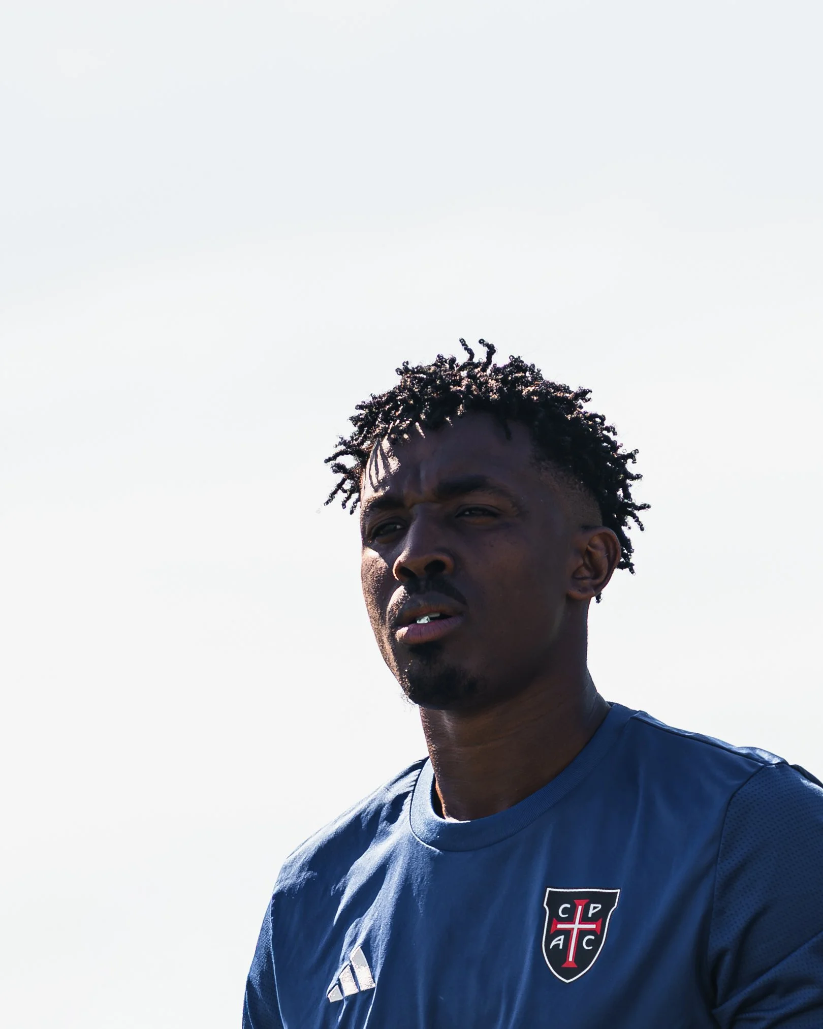 A young man with dark skin and short curly hair is wearing a blue sports jersey with a crest on it. The background is clear and bright sky.