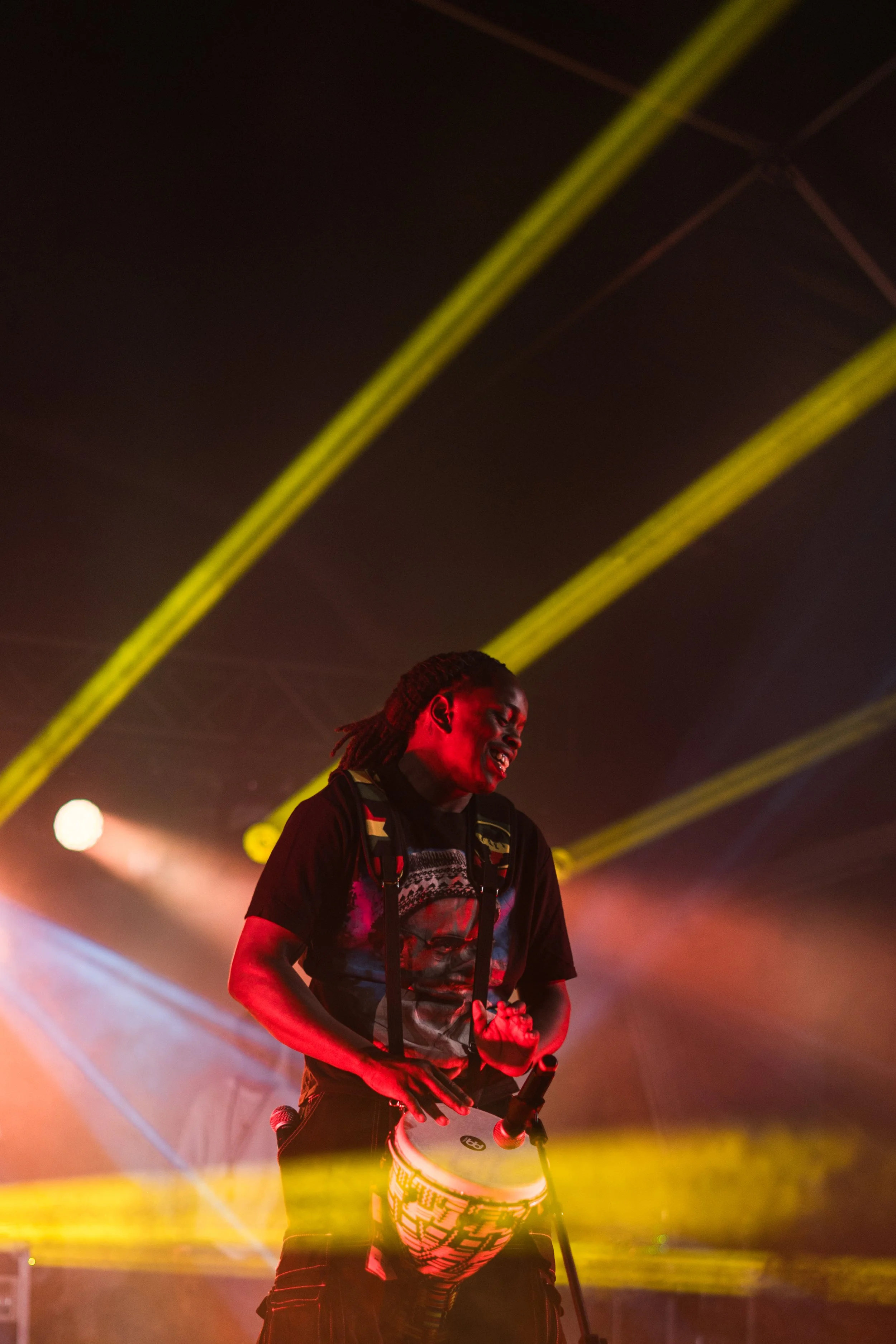 A person with dreadlocks playing drums on stage, illuminated by warm stage lights and yellow beams.