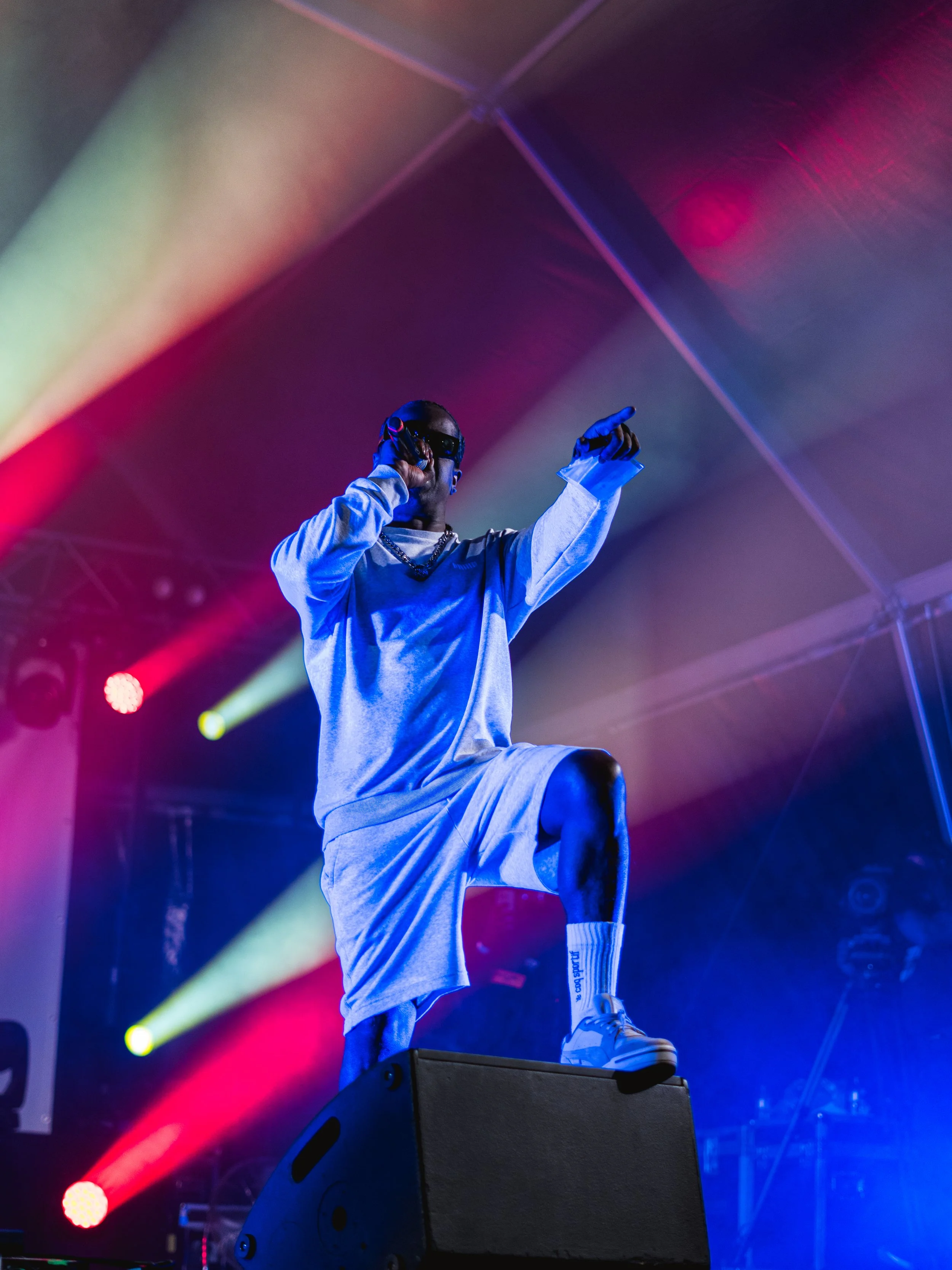Performer on stage holding a microphone and pointing with the other hand, wearing sunglasses, a long-sleeve shirt, shorts, and sneakers, with colorful stage lighting and confetti in the background.