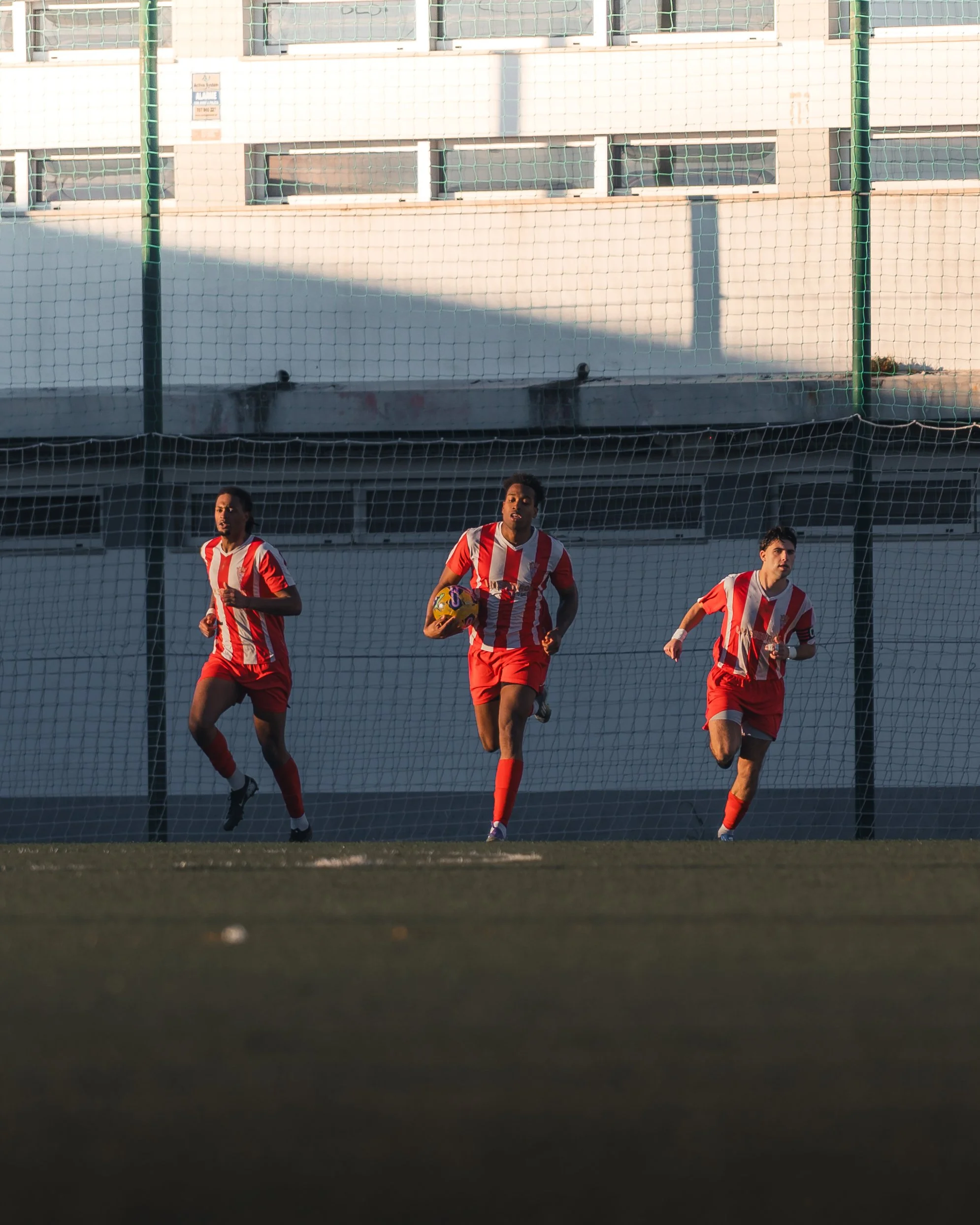 Football players in red and white uniforms running on a field behind a net, with a building in the background.