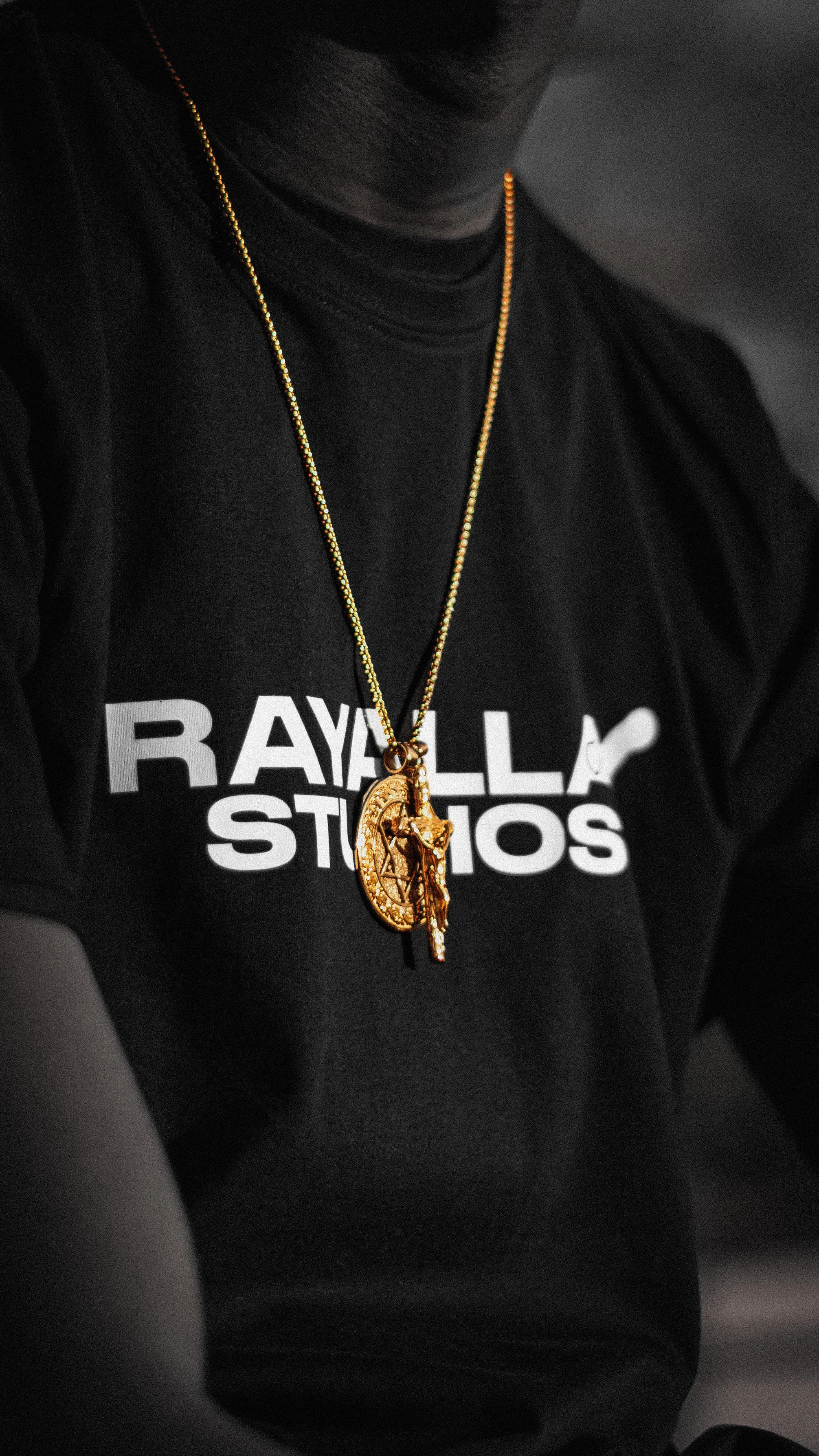 Close-up of a person wearing a black T-shirt with white text and gold jewelry, including a chain with religious pendants, against a dark background.