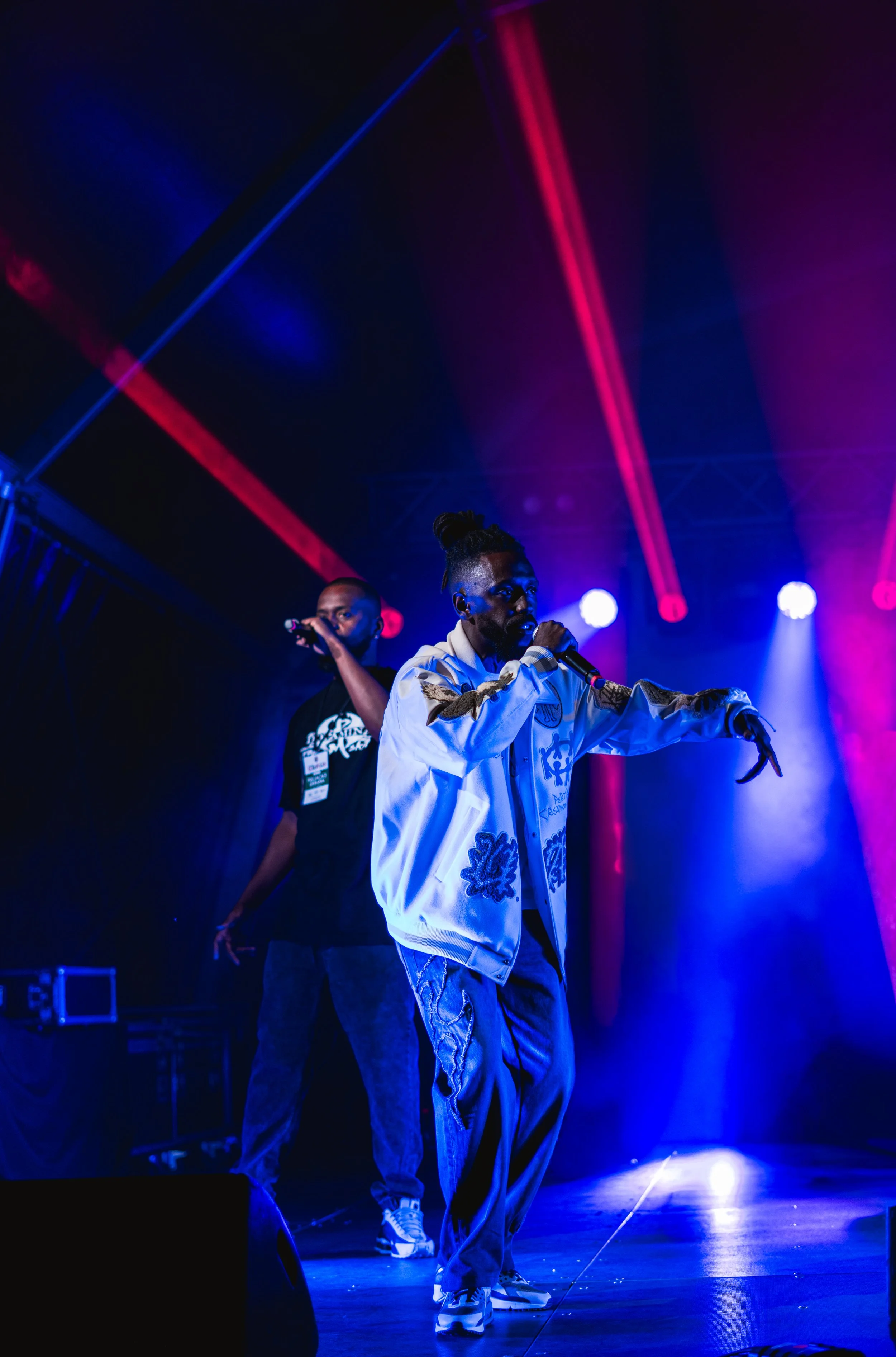 Two men on stage under colorful stage lighting, one with a microphone and the other standing with his hands in his pockets, performing at a show
