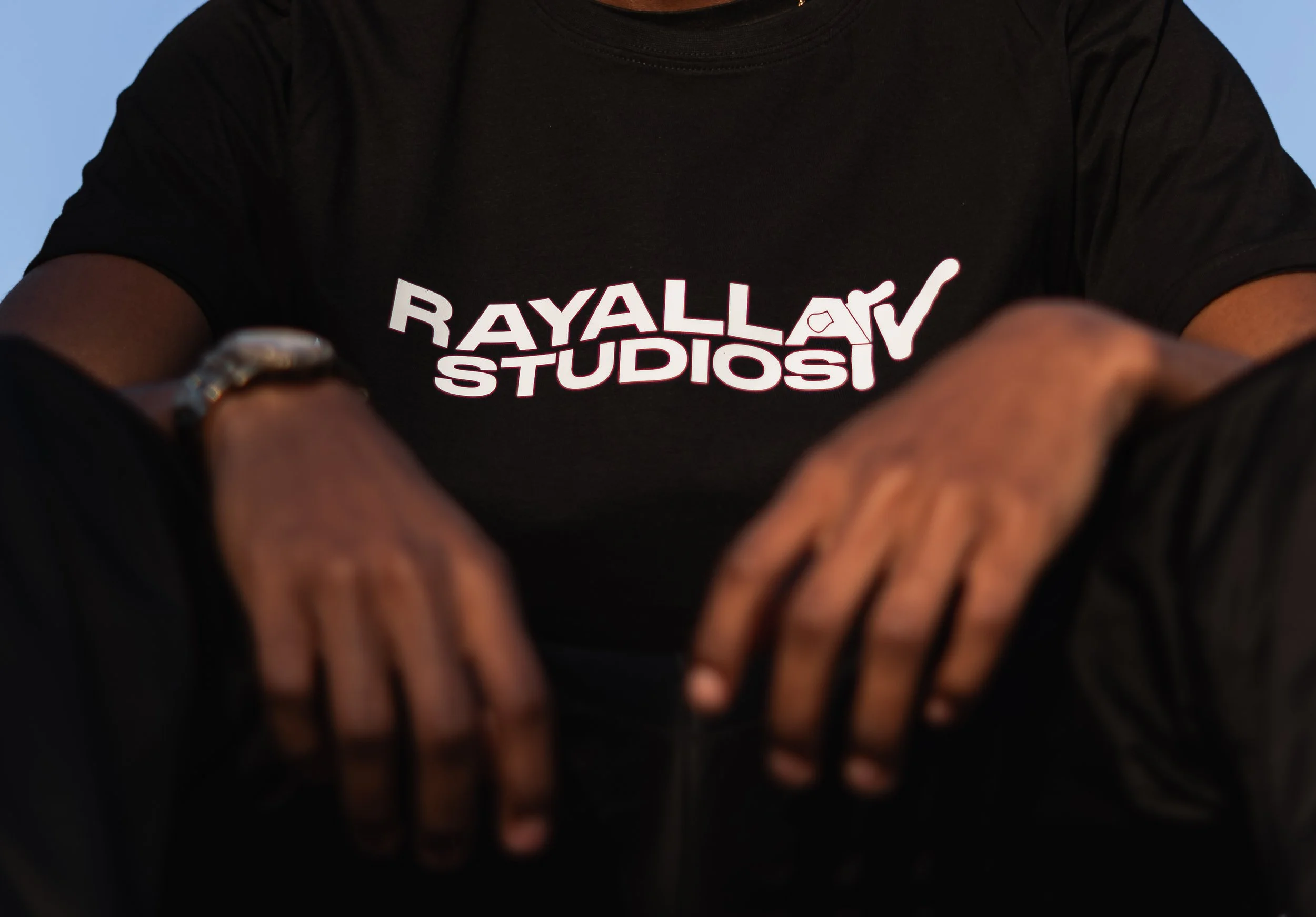 Person wearing a black T-shirt with a logo and text that reads 'RAYALLA STUDIOS'. The person is sitting, wearing a watch on the left wrist and a ring on the right hand.