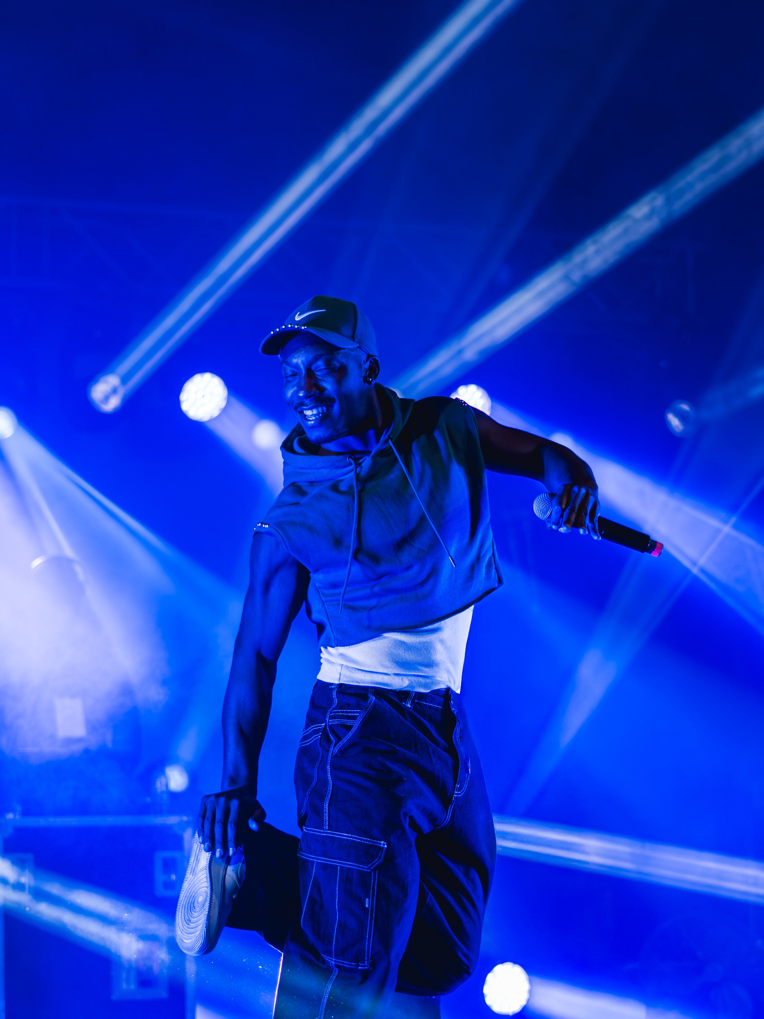 A male performer on stage during a concert, wearing a Nike hoodie and baseball cap, holding a microphone with bright stage lights and blue lighting in the background.