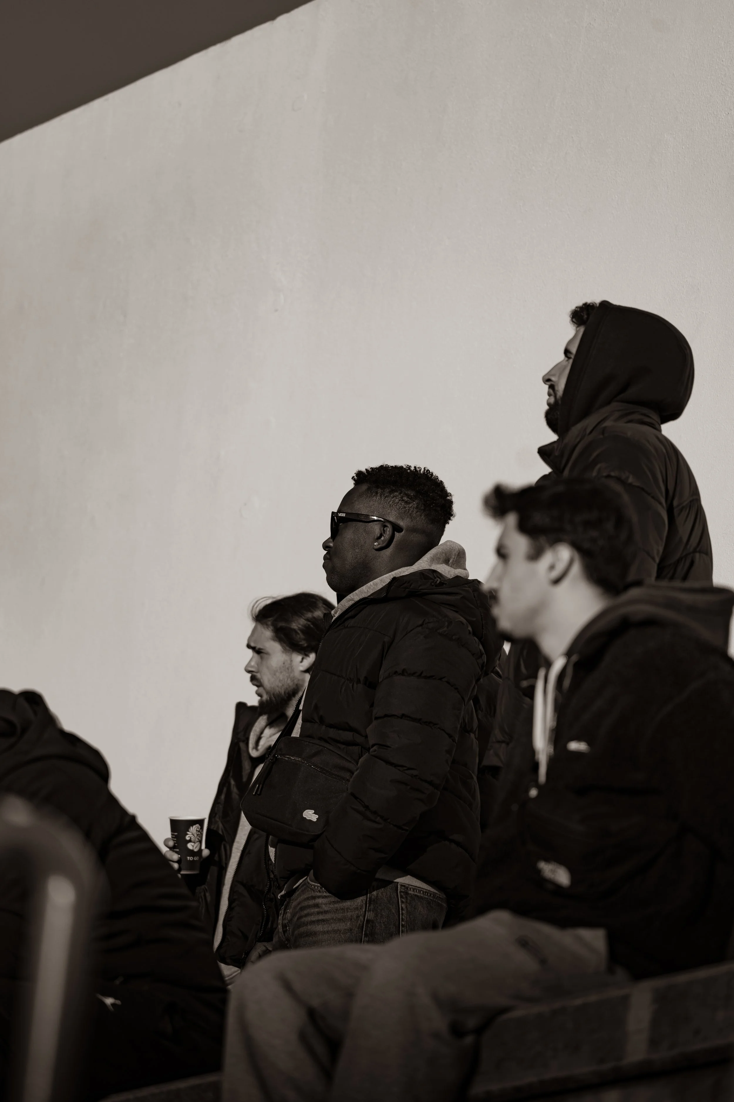 A group of men standing in the stands, wearing jackets, some with hoodies, in front of a plain wall, with one person holding a cup.