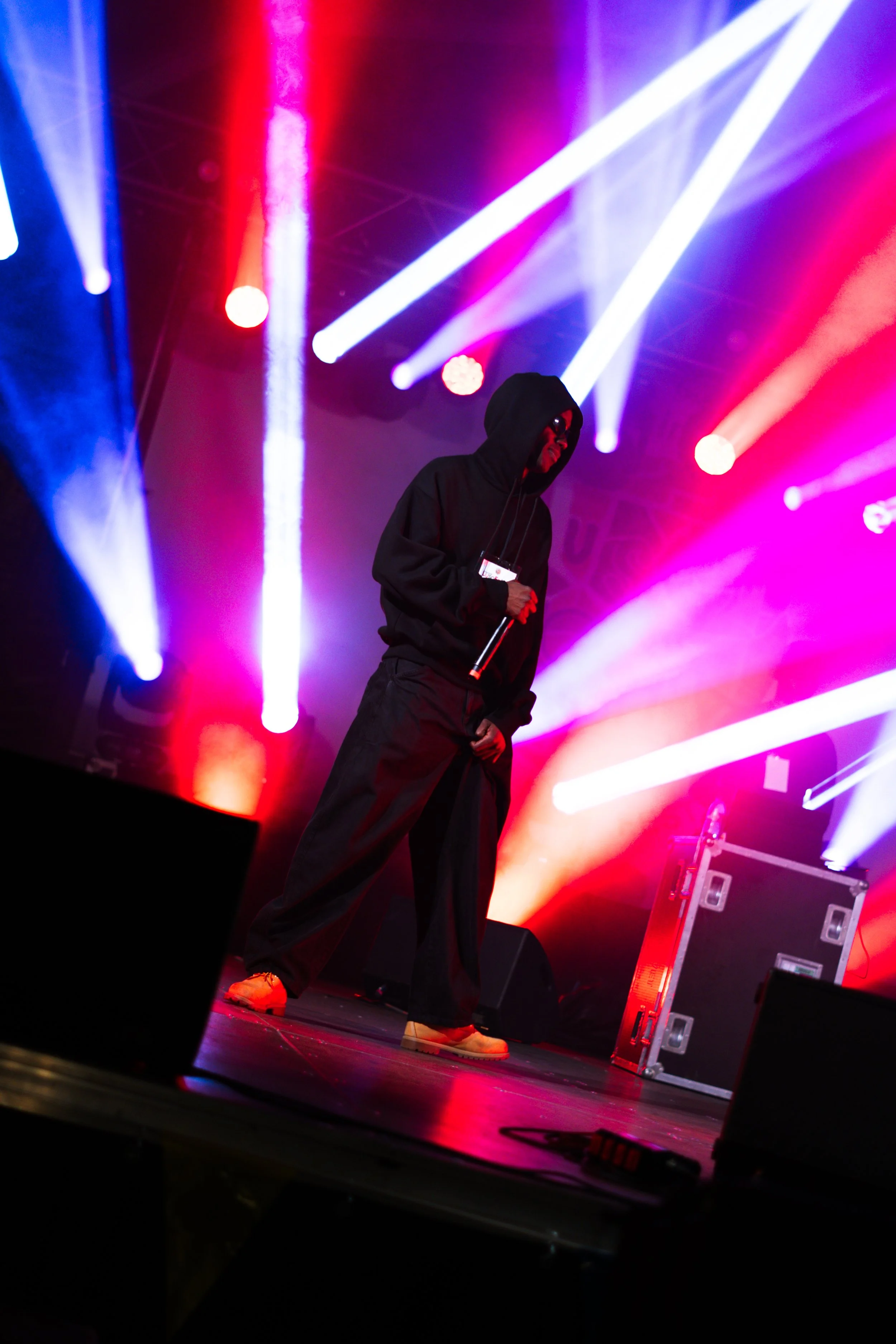 A performer on stage wearing a black hoodie, dark wide-leg pants, and beige shoes, holding a microphone, under colorful stage lights with red, purple, white, and blue beams.