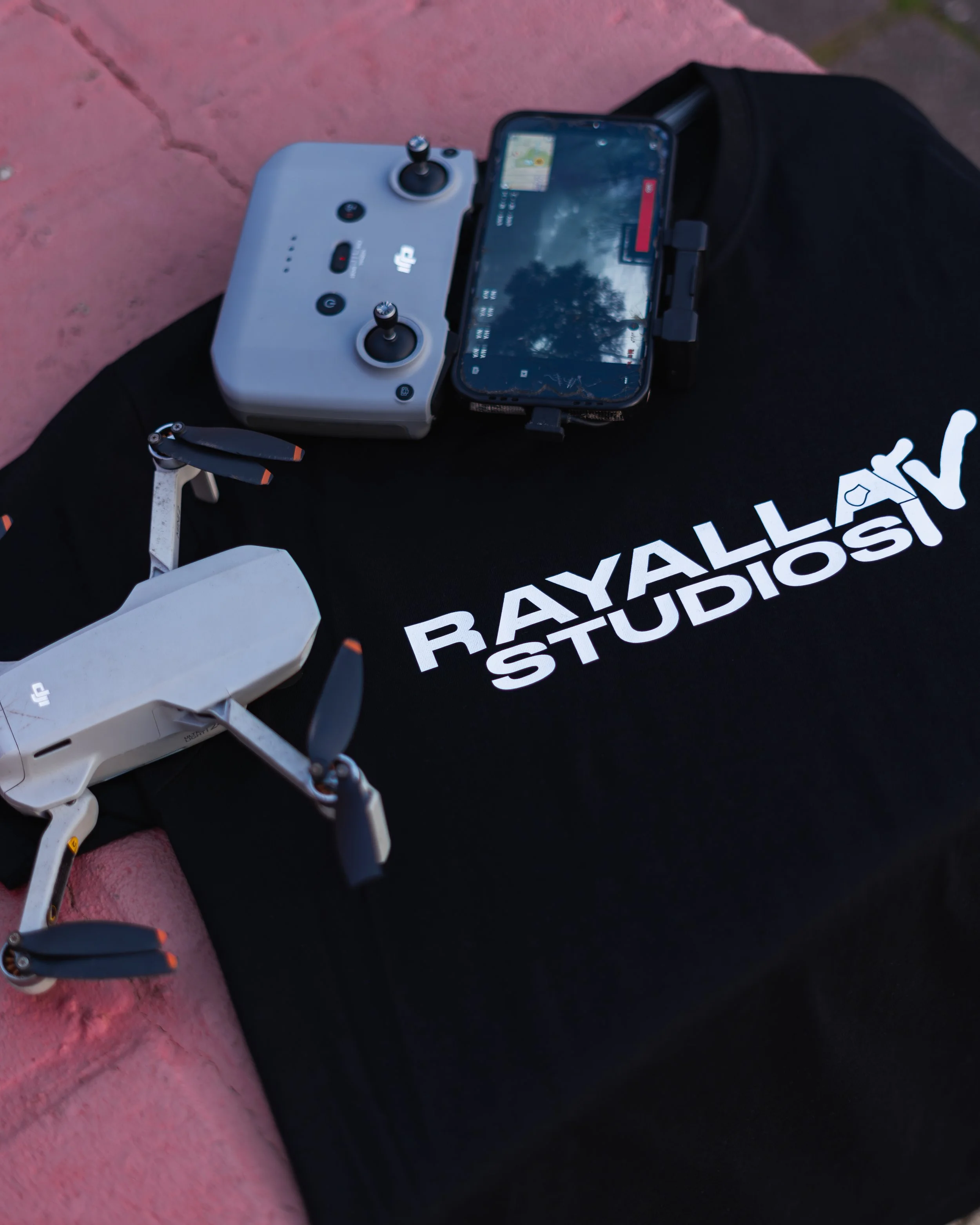 A drone remote controller with a smartphone mounted on it, placed on a black shirt with white text, next to a pinkish surface. The smartphone has a visible screen showing a flight interface.