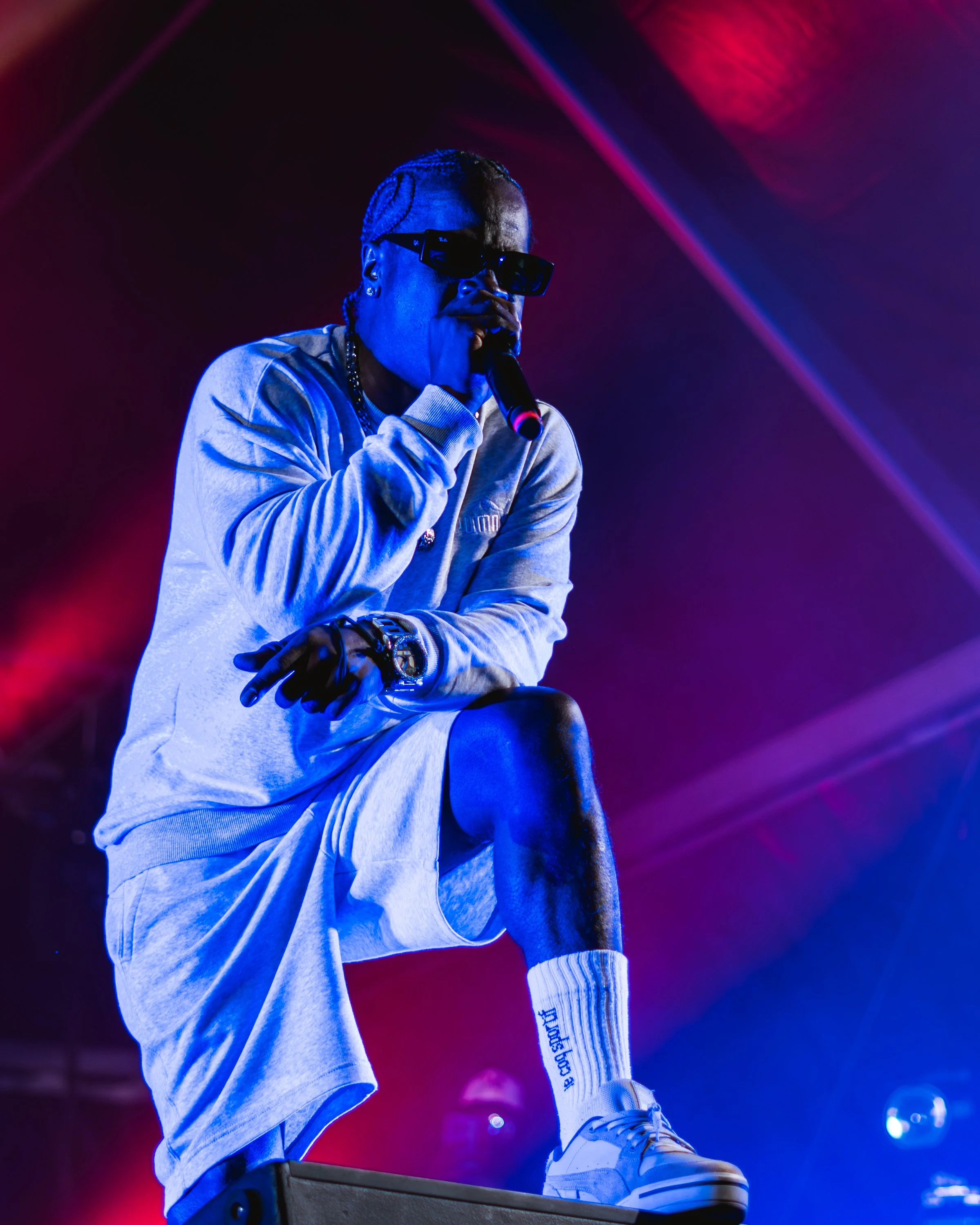 A rapper performing on stage, wearing sunglasses, a light gray sweatshirt, white shorts, a chain necklace, and white sneakers, with colorful stage lighting in the background.