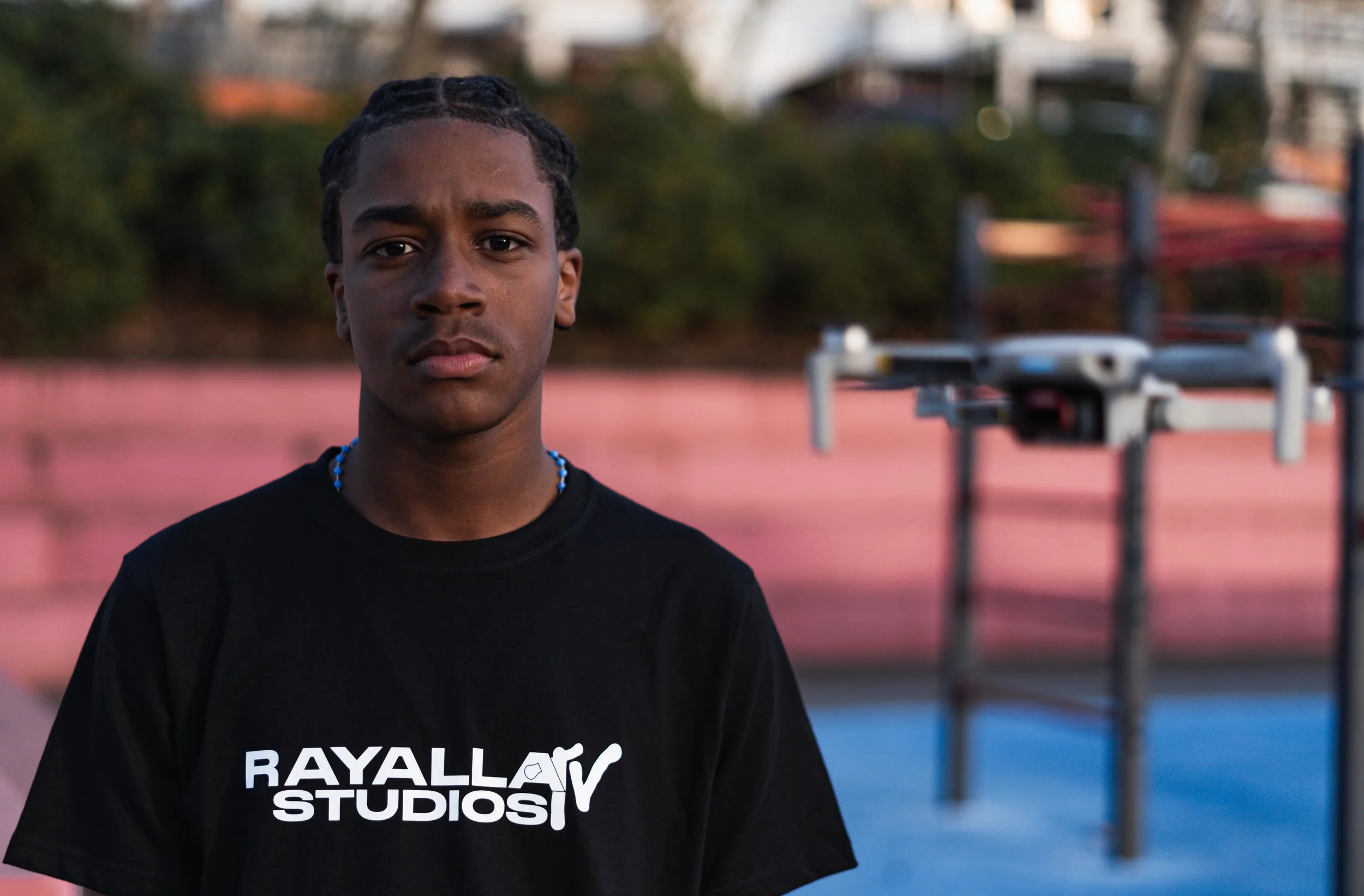 A young man with braided hair wearing a black T-shirt that says 'RAYALLA STUDIOS' stands outdoors on a colorful sports court with blurred background of trees and buildings.