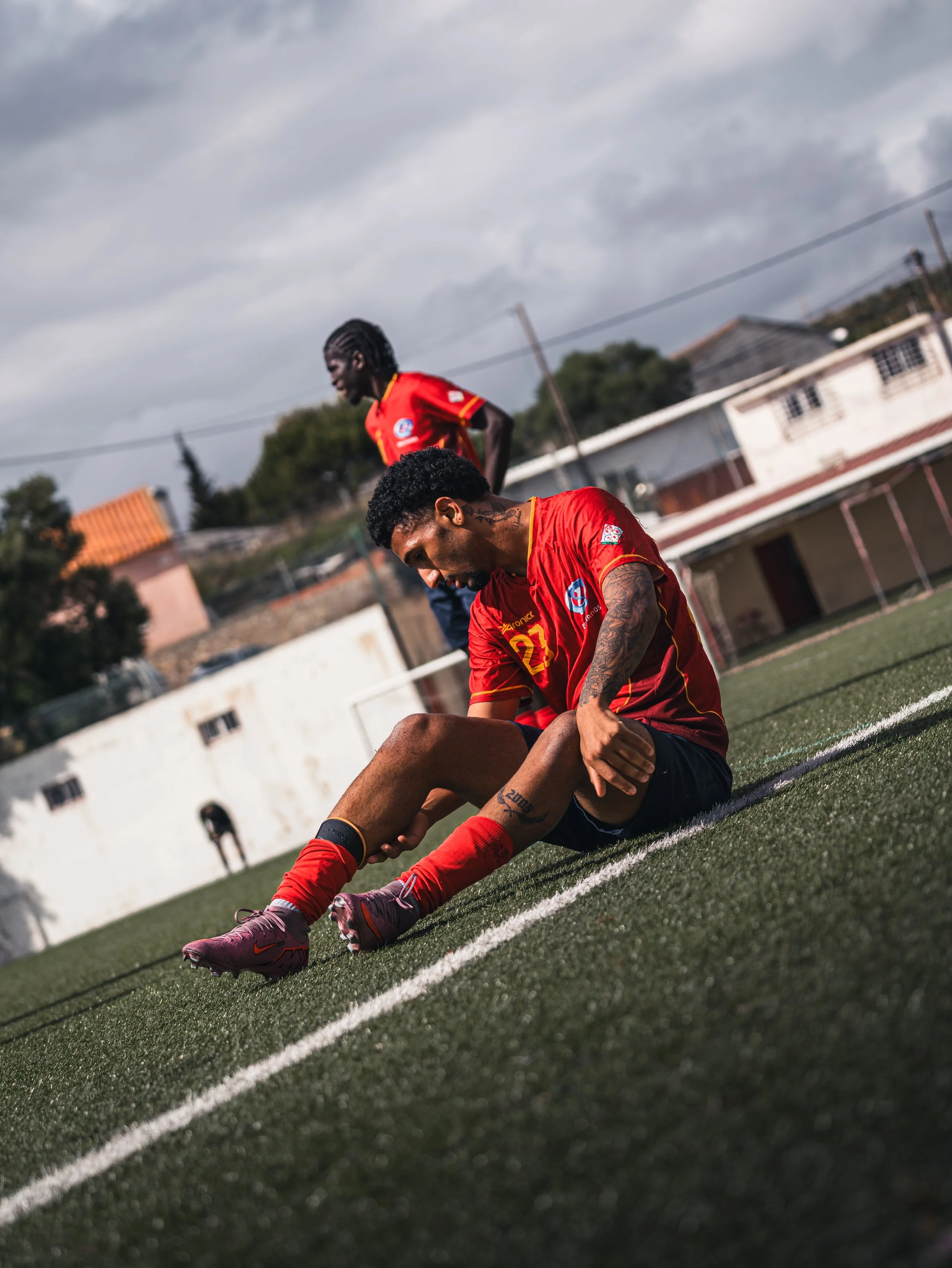 Two football players in red uniforms on the field, with one sitting on the ground and the other standing behind him, under a cloudy sky.