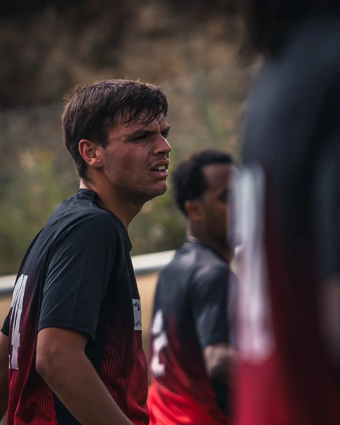 A young man with wet, dark hair and a serious expression, wearing a black and red sports jersey, standing outdoors during a sporting event.