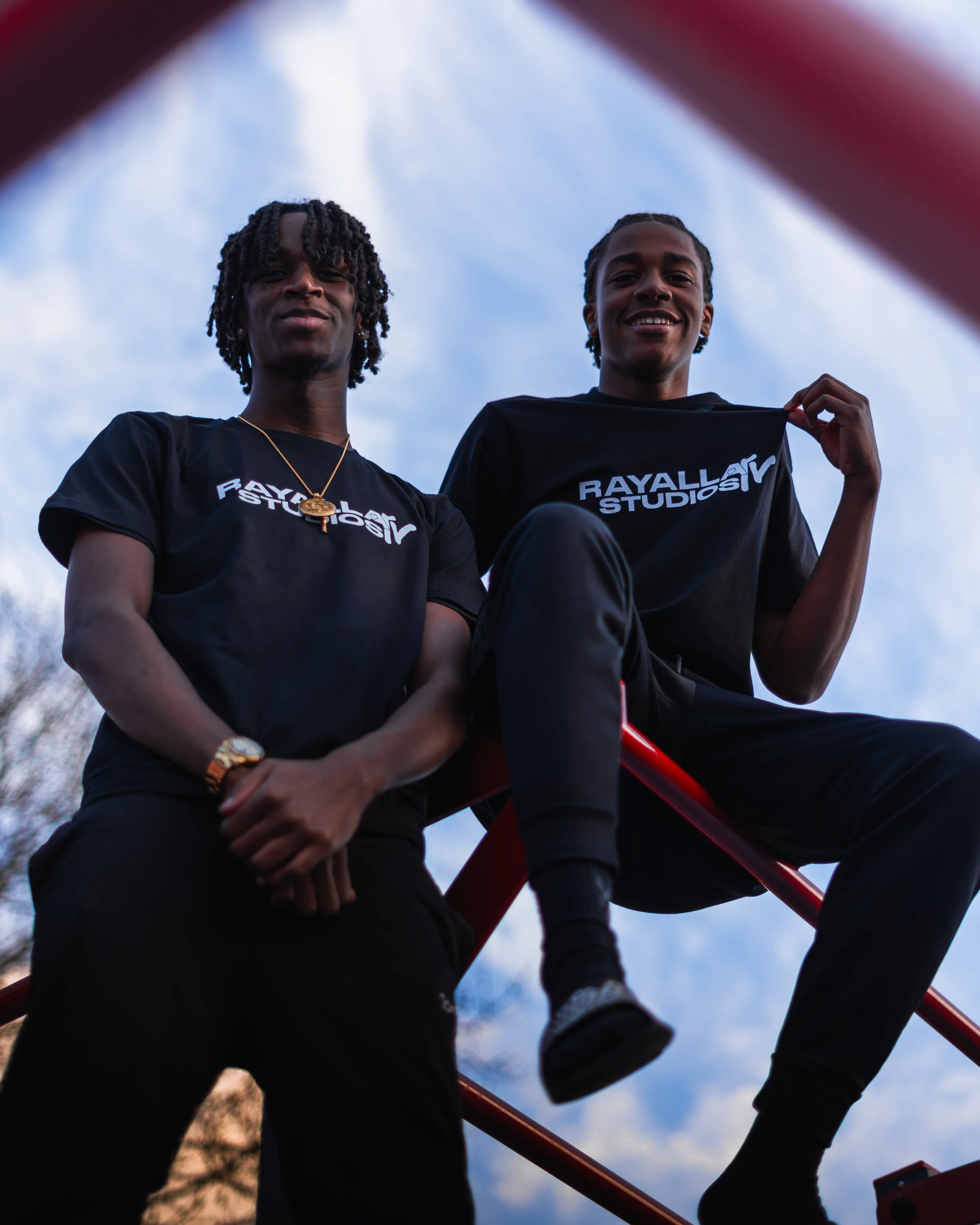 Two young men sitting on a red playground structure, looking down at the camera and smiling, with a blue sky and clouds in the background.