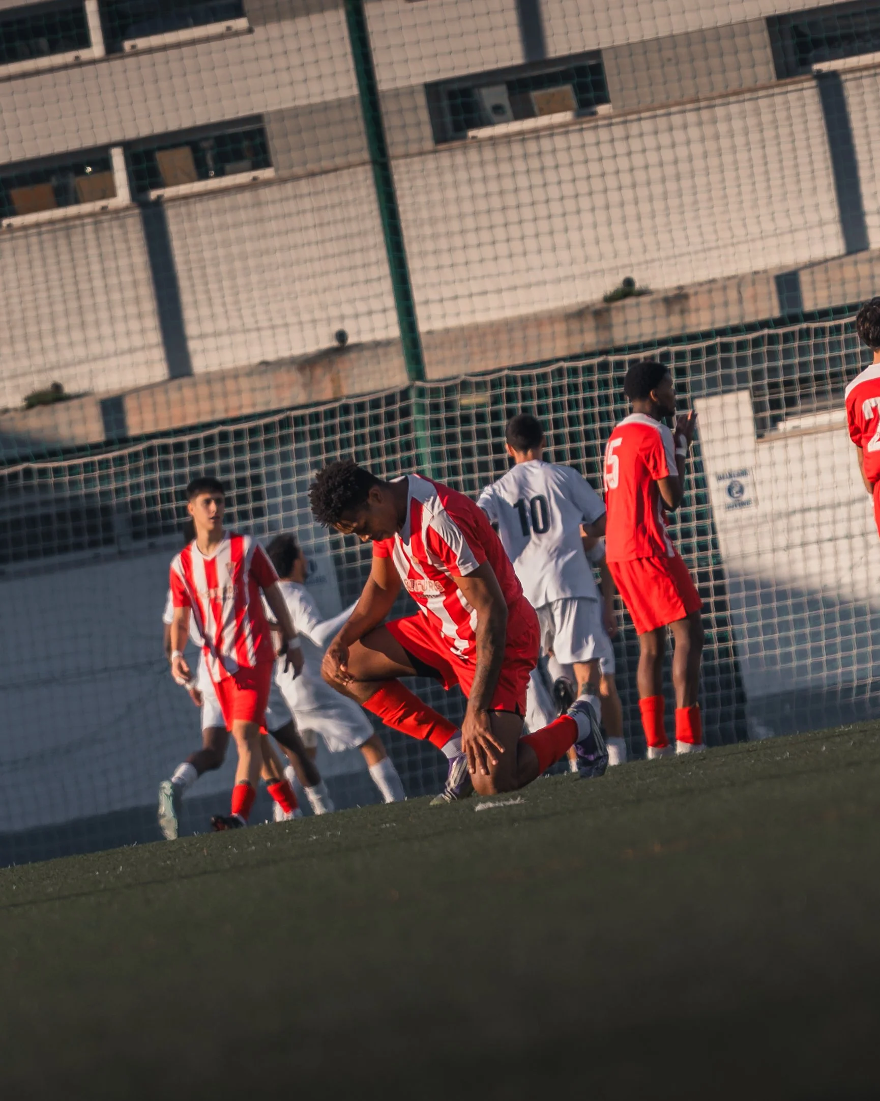 Football players in red and white uniforms on the field during a game, with a netted fence and residential buildings in the background.