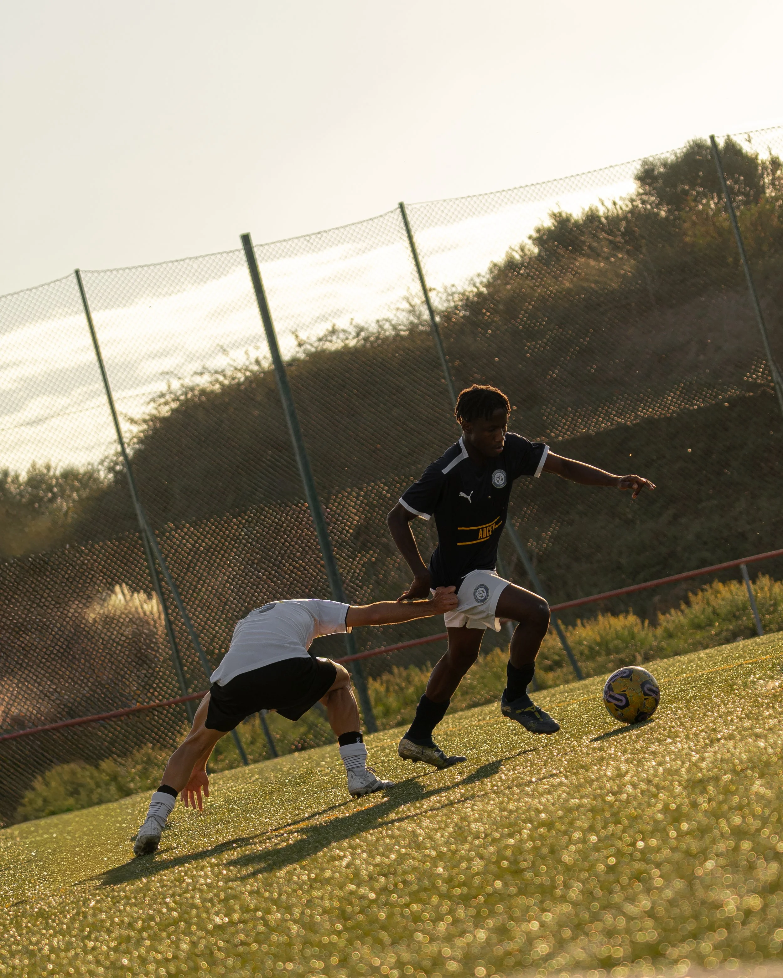 Two football players are on a field during sunset, with one player leaning forward and the other running with the ball, near a chain-link fence and a hillside with trees in the background.