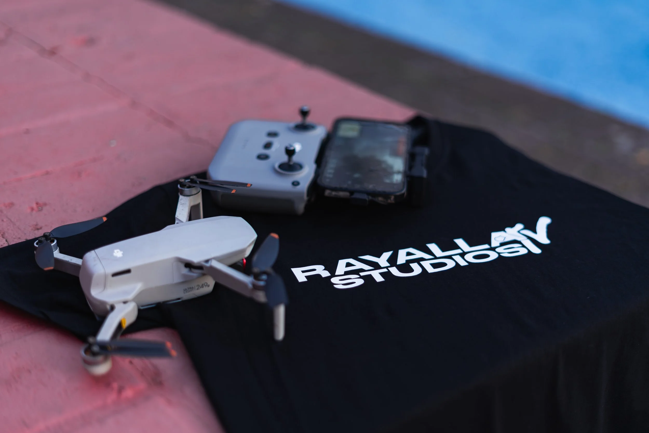 A drone with white body and black propellers resting on a black cloth with white text. Next to the drone, there is a remote control device with joysticks and a smartphone mounted on top. The background shows a surface painted pink with a blue section