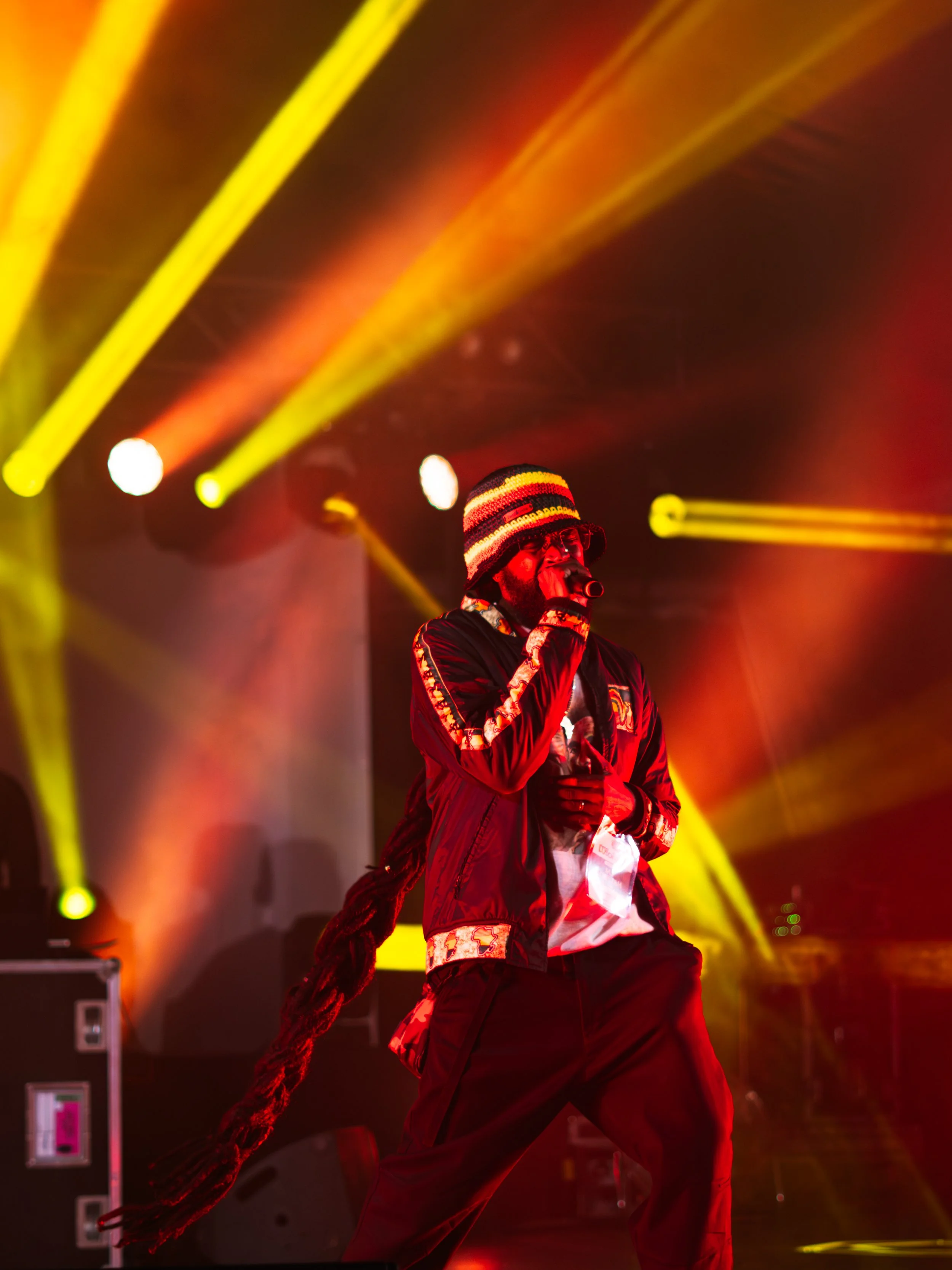 A male performer wearing a striped knit hat, glasses, and a jacket, singing into a microphone on stage under red, yellow, and orange stage lights.