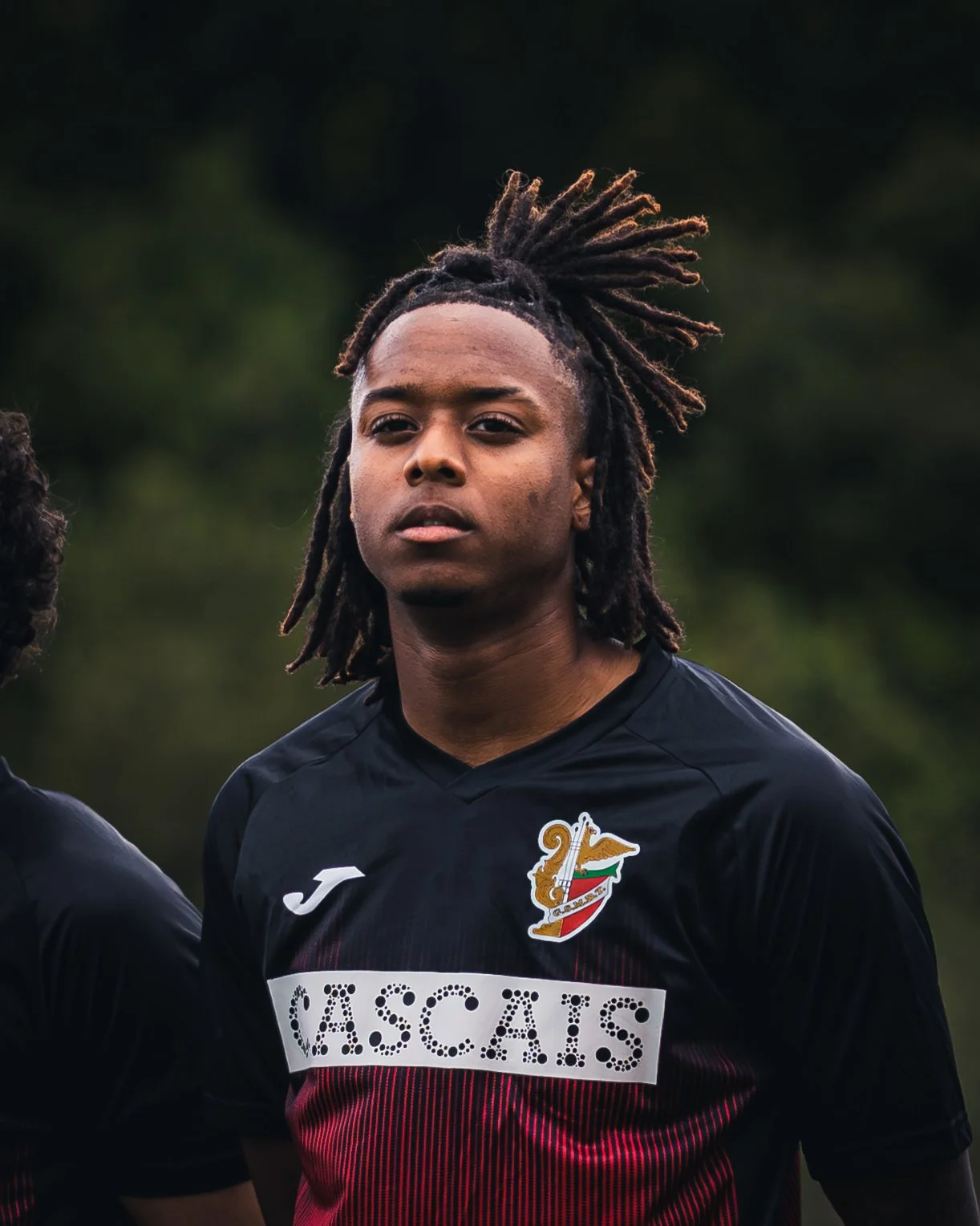 A young man with dreadlocks wearing a black soccer jersey, looking into the distance outdoors with a blurred green background.