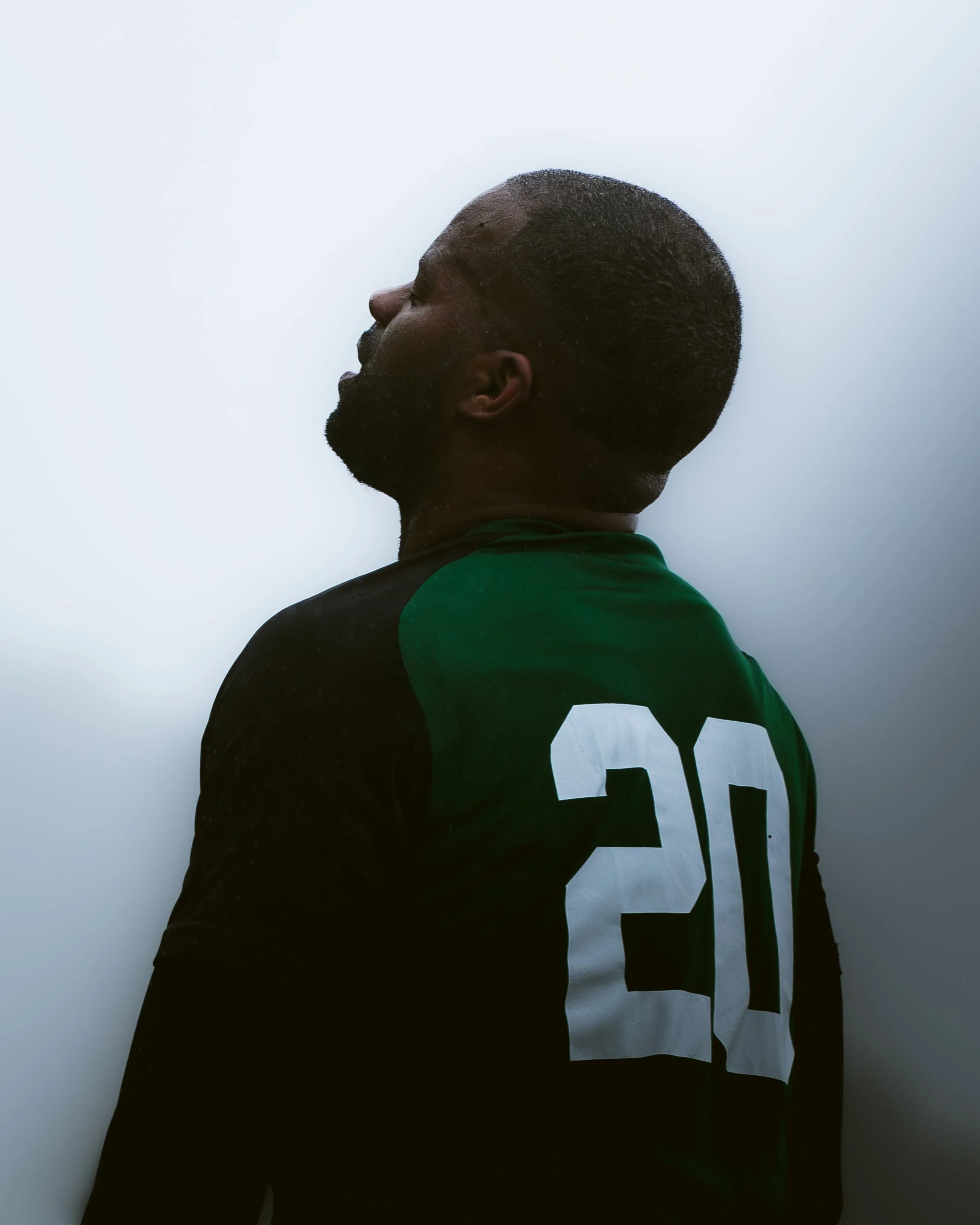 Side profile of a man with a trimmed beard, wearing a black and green shirt with white graphic letters, against a plain light background.