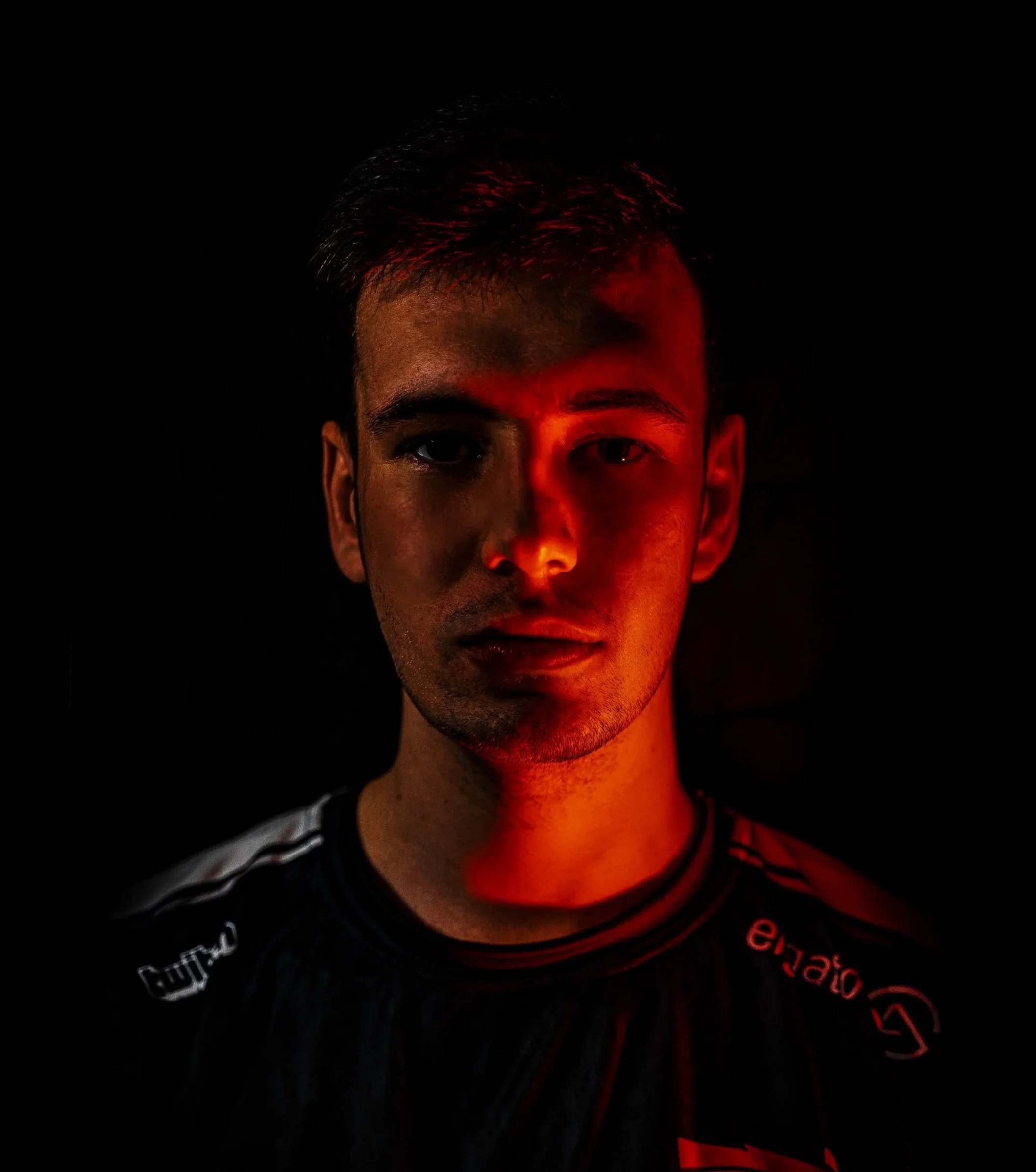 Close-up of a young man's face illuminated by red light, dark background, wearing a black shirt with red and white text.